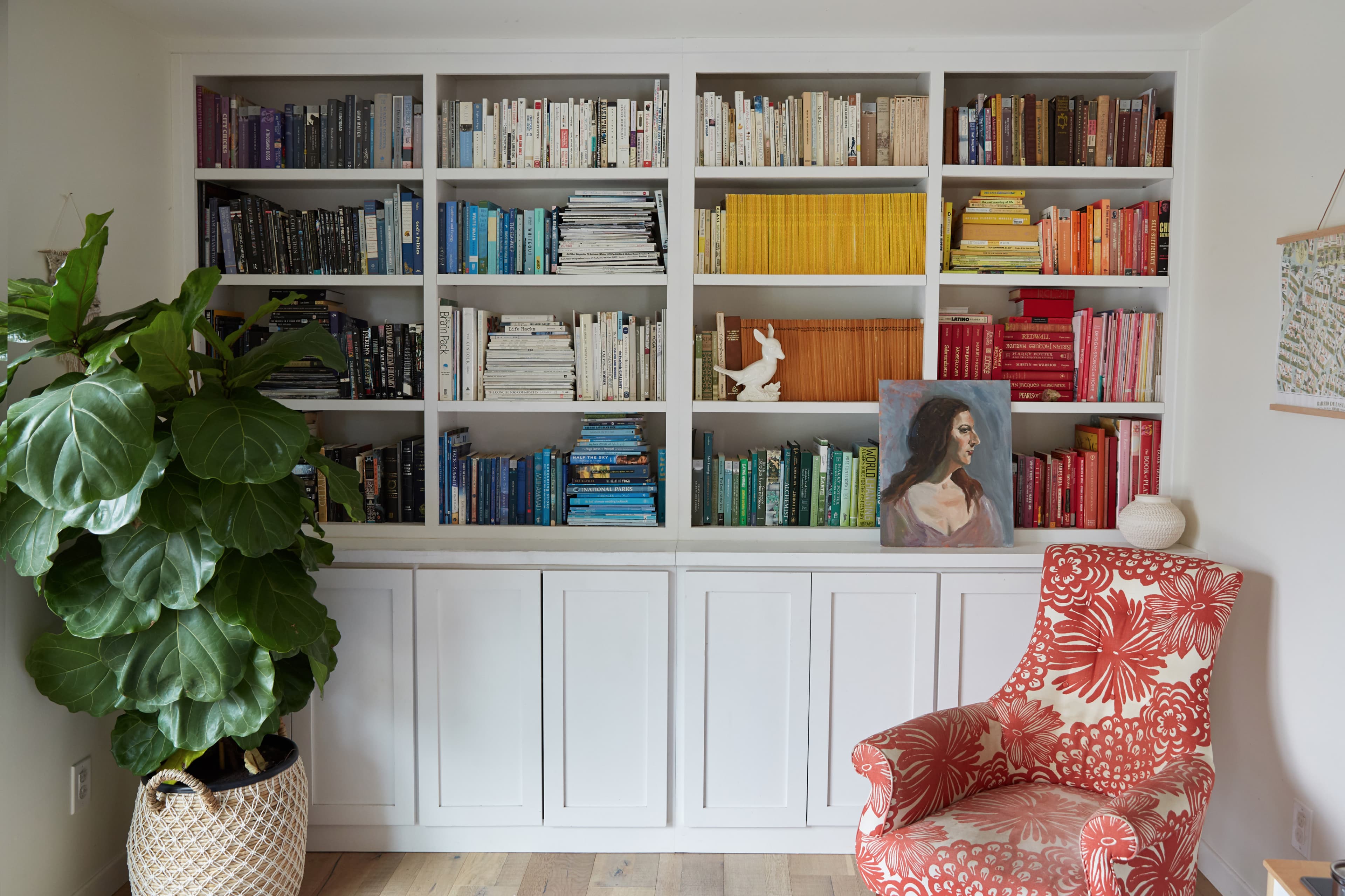 A bookcase filled with neatly arranged books of various colors, accompanied by a potted plant, a chair with a floral pattern, and a painting of a woman.
