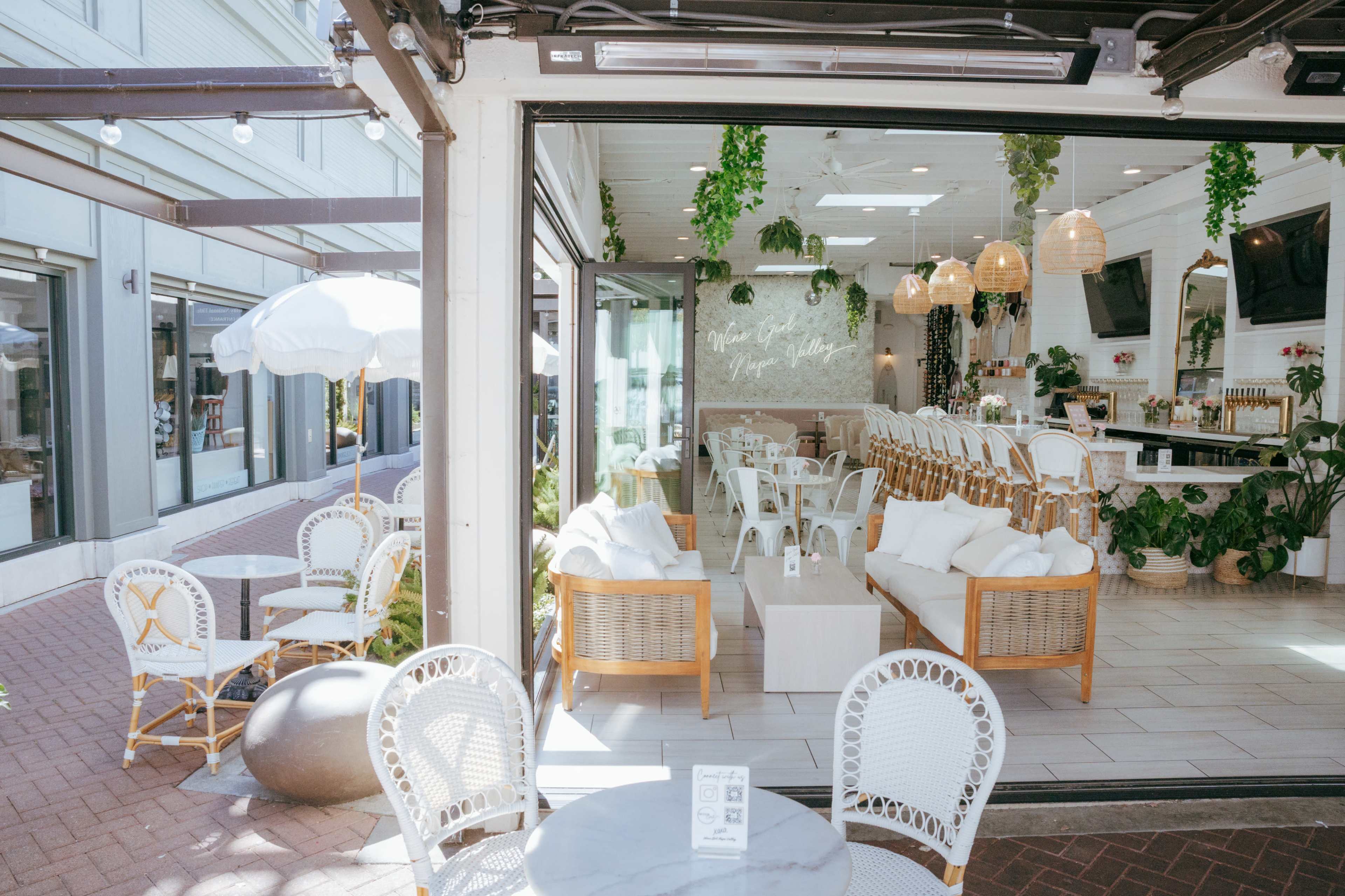 A bright café features wicker furniture, large windows, and greenery, creating an inviting atmosphere for guests.