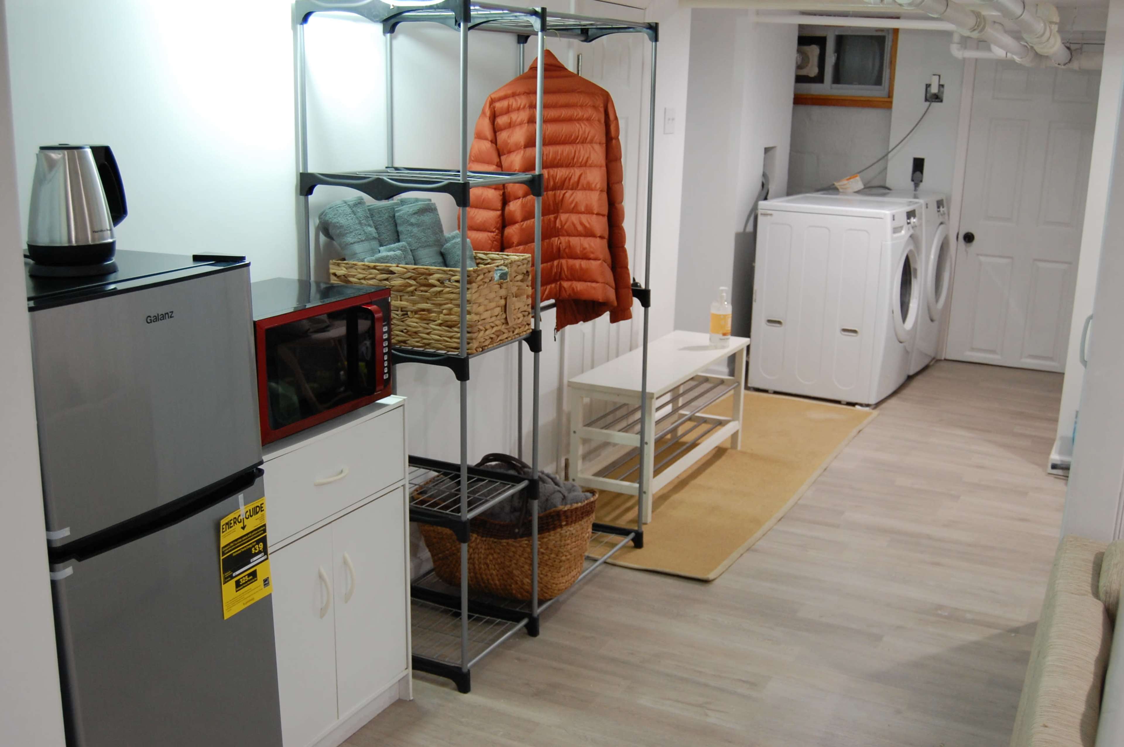 The image shows a domestic laundry area featuring a stacked washer and dryer, a small refrigerator, a microwave, shelving with various items, and a tan rug on the floor.