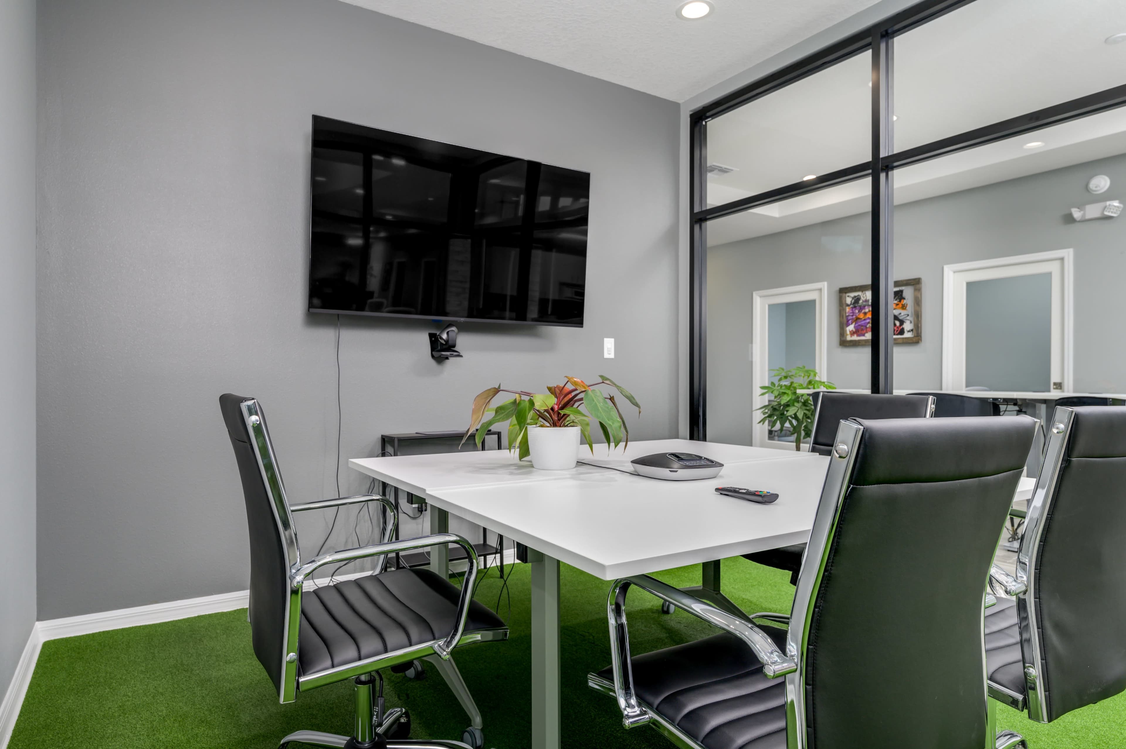 A conference room features a modern table with four black chairs, a wall-mounted TV, and potted plants, all set against a gray backdrop.