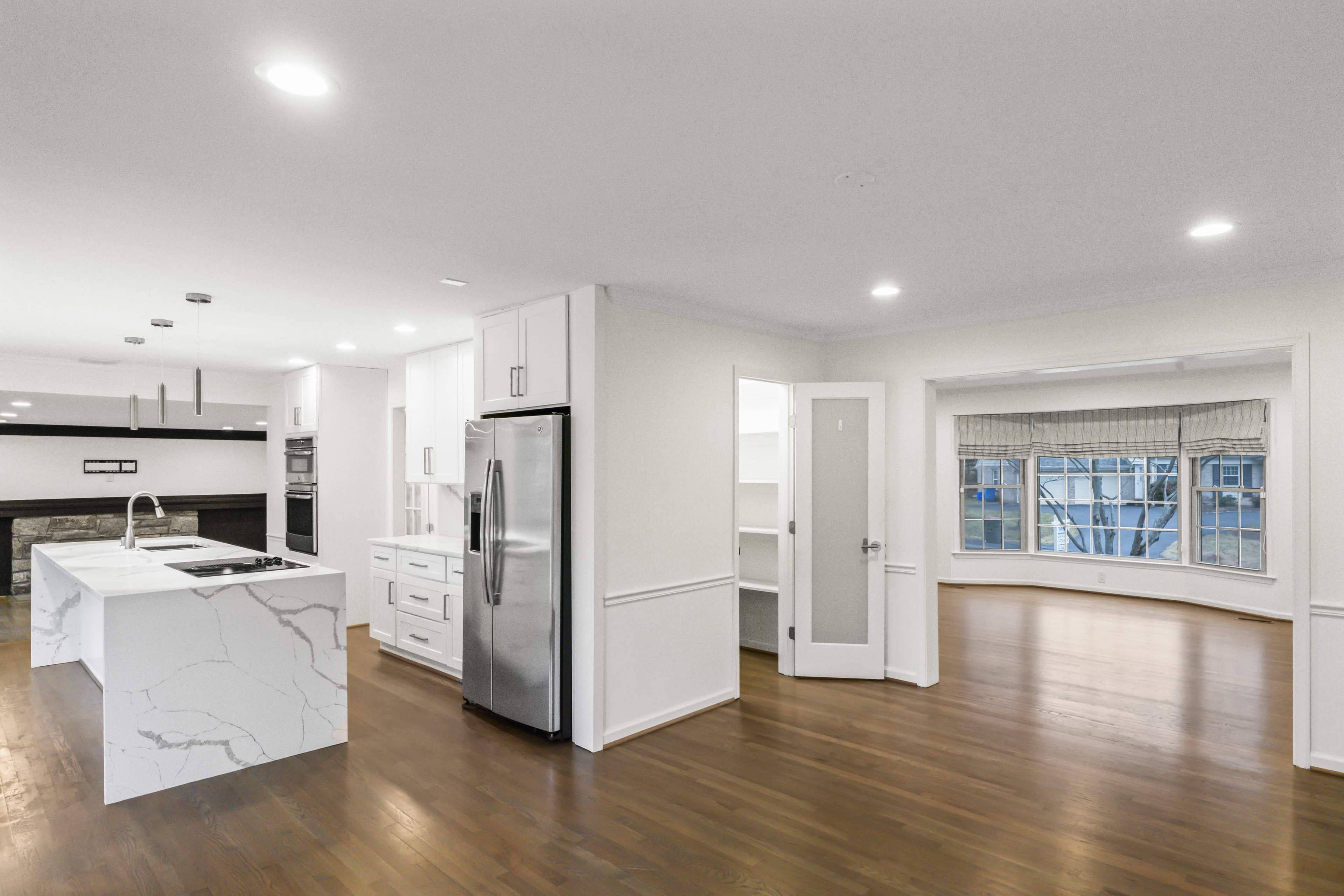 The image shows a modern kitchen and living area with hardwood floors, featuring a marble countertop island, stainless steel appliances, and large windows.