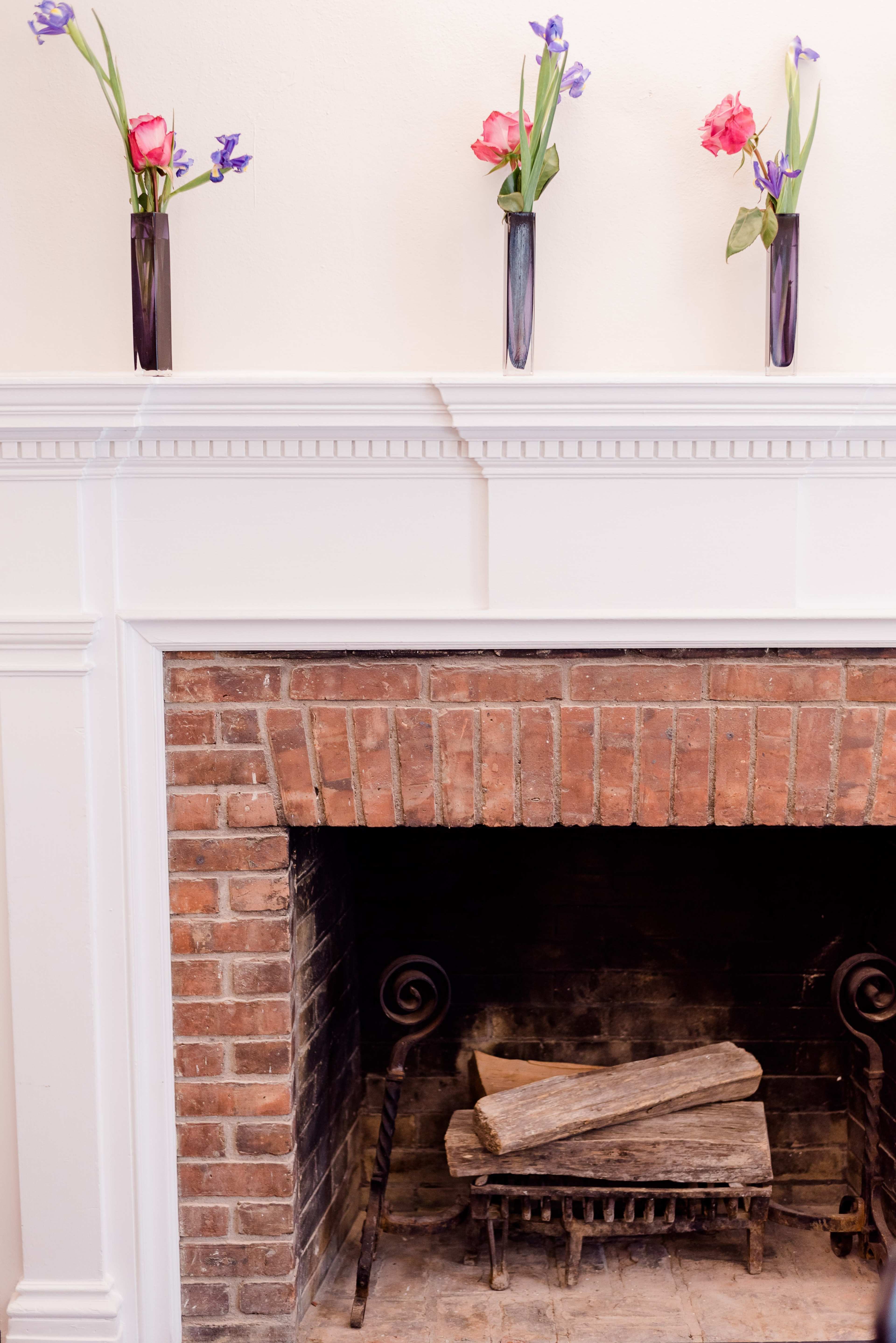 A brick fireplace with a wood log holder contains stacked logs, topped by a white mantel displaying three vases of flowers.