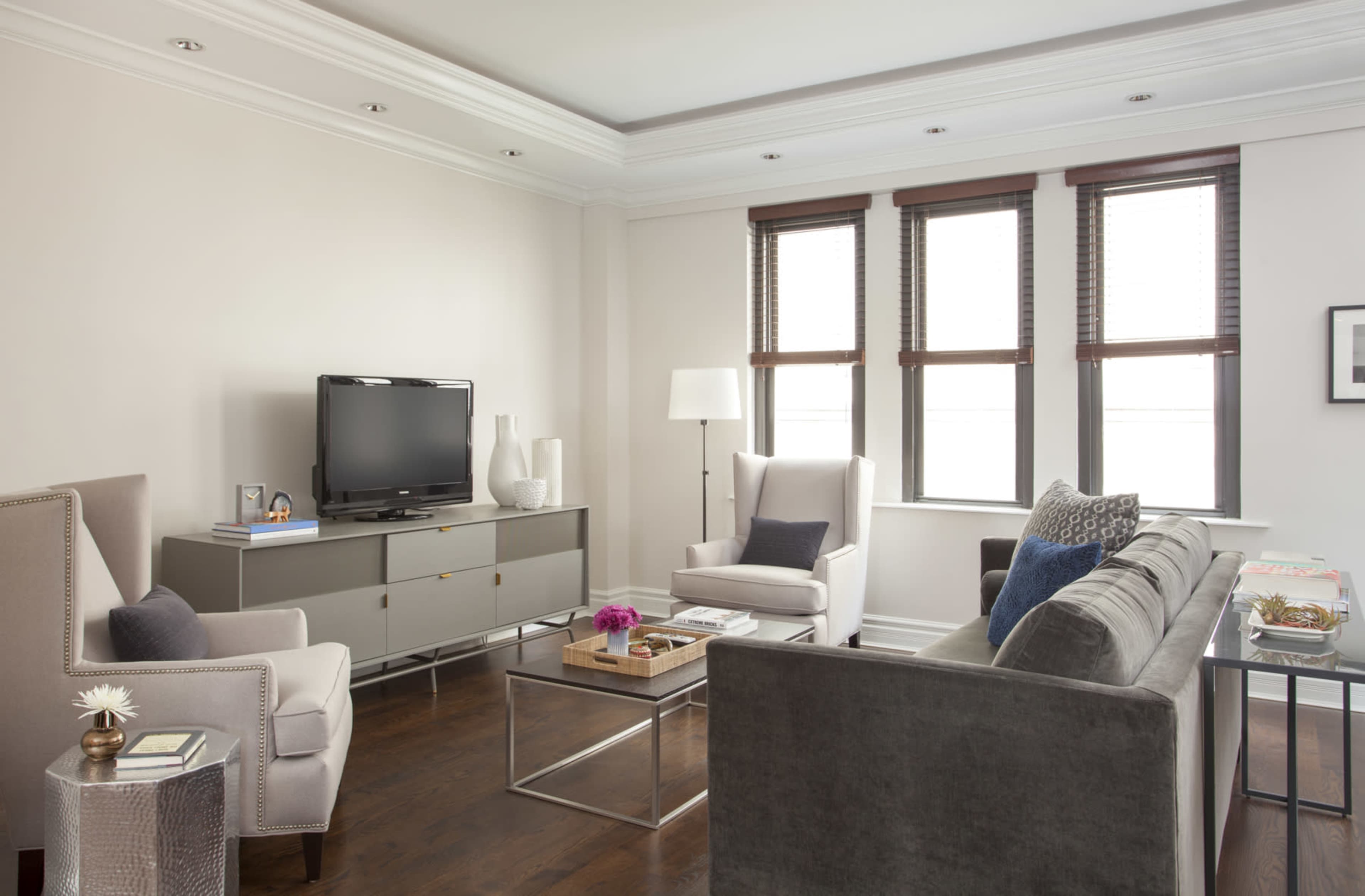 A modern living room features a gray sofa, two armchairs, a television on a media console, and three windows with blinds, illuminated by natural light.