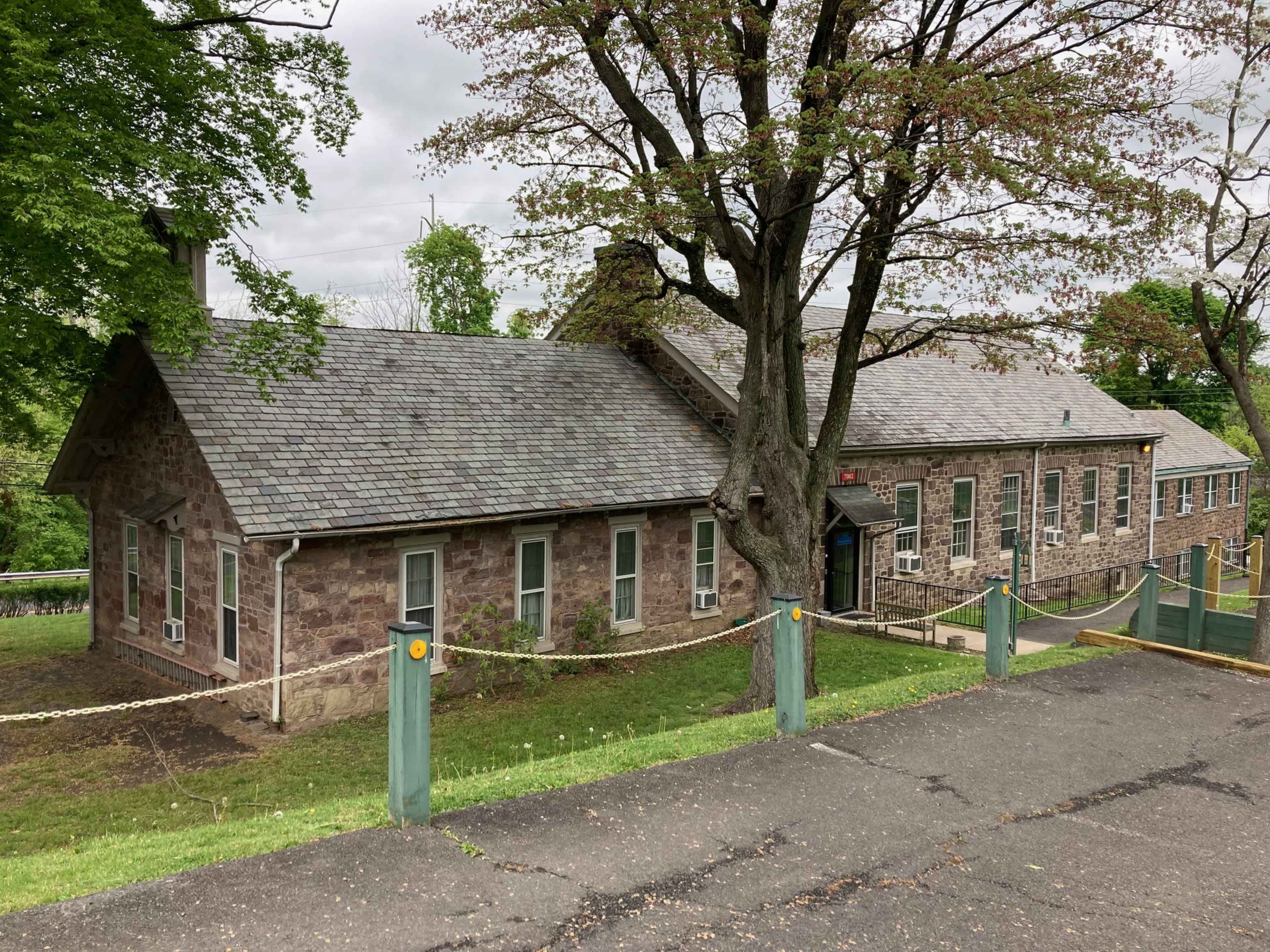 The image shows a stone building with a sloped roof, surrounded by green grass and trees, located on a hillside.