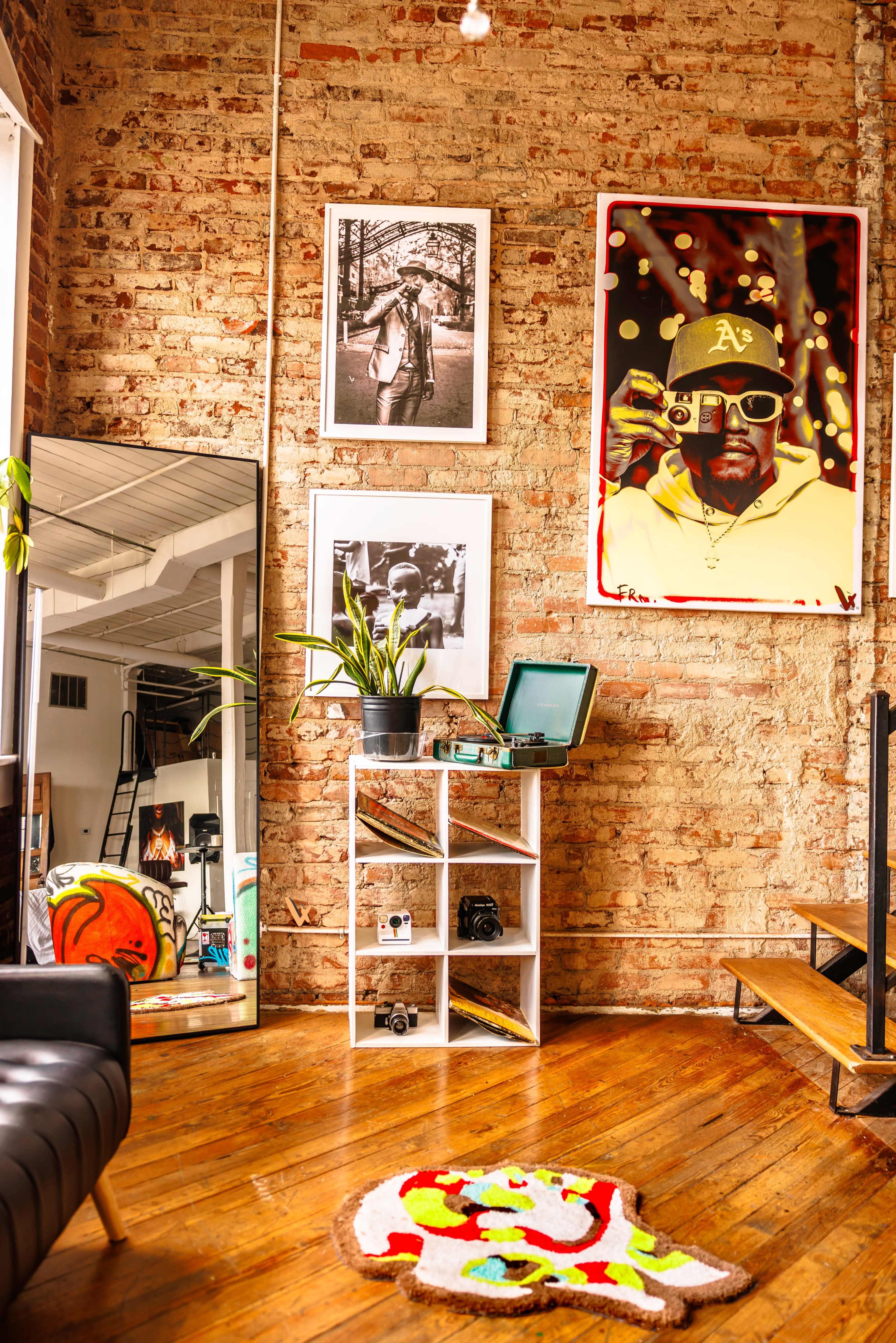 The image shows a cozy interior space featuring exposed brick walls, framed photographs, a large colorful portrait, a potted plant, a vintage record player on a shelf, and a multicolored rug on a wooden floor.