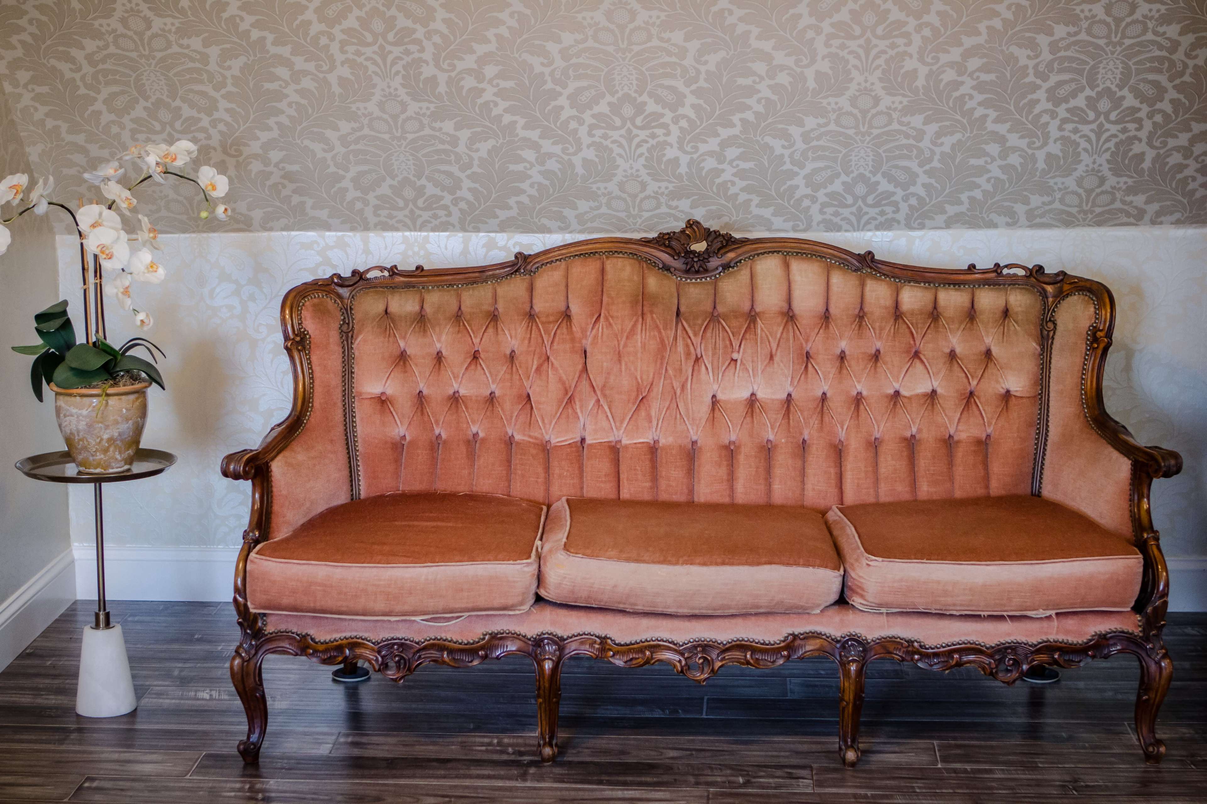A vintage-style upholstered couch with an ornate wooden frame, accompanied by a small side table holding a potted plant with white flowers.