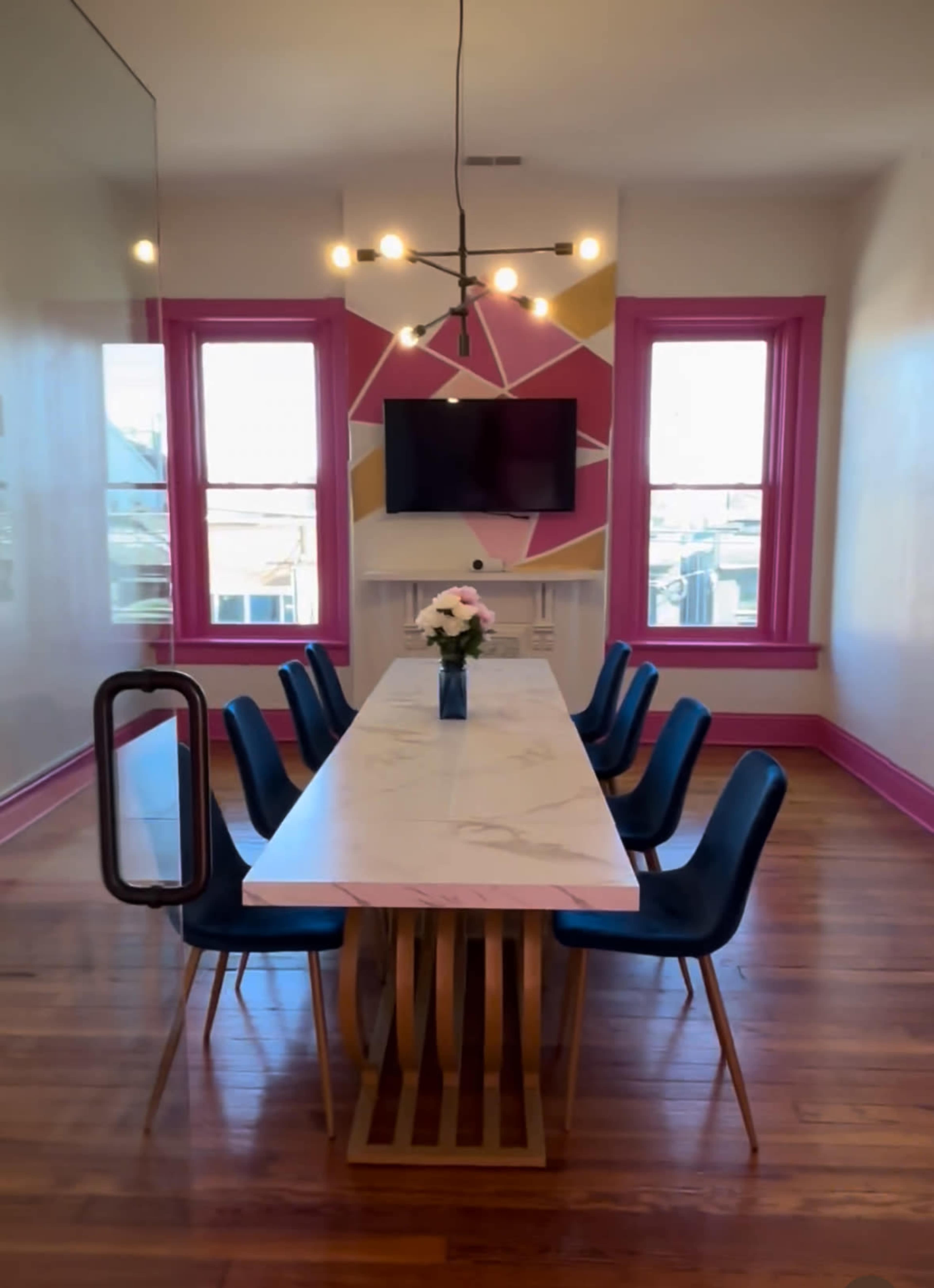 A spacious conference room features a long marble table surrounded by six black chairs, with modern lighting and a colorful accent wall in pink and yellow.