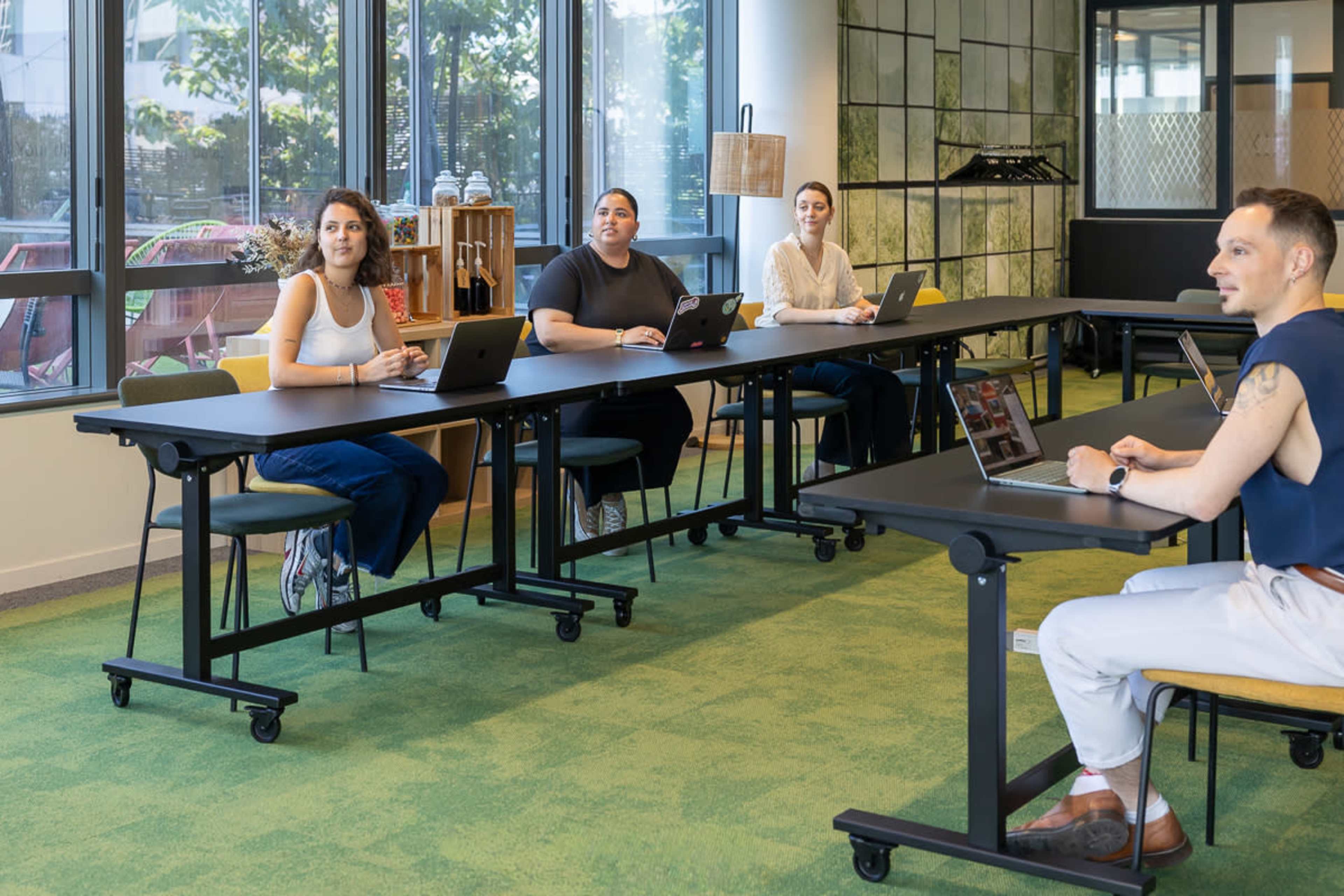 Four people sit at a long table in a modern meeting room, each using a laptop and facing a speaker.