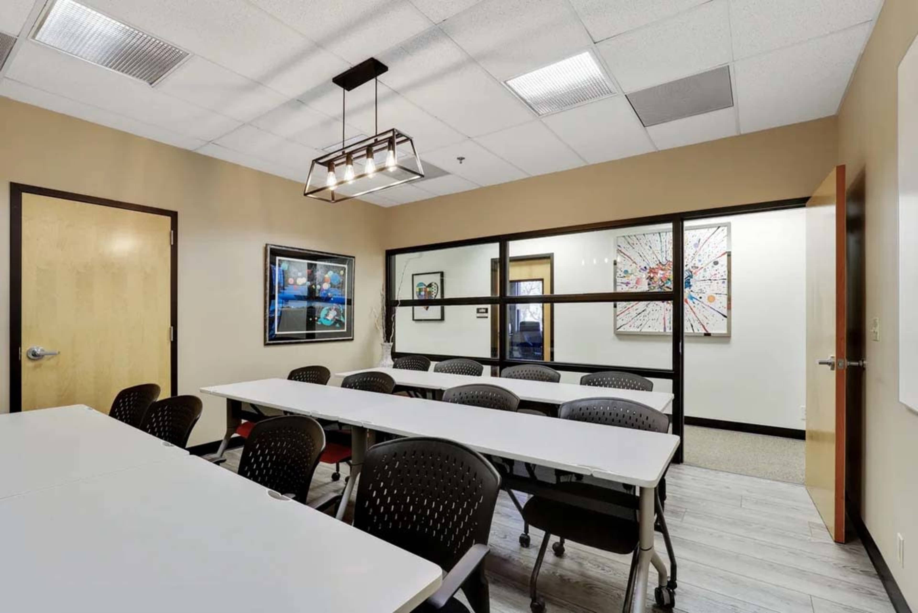 The image shows a modern conference room with rows of tables and black chairs, featuring framed artwork and large windows that create a bright atmosphere.