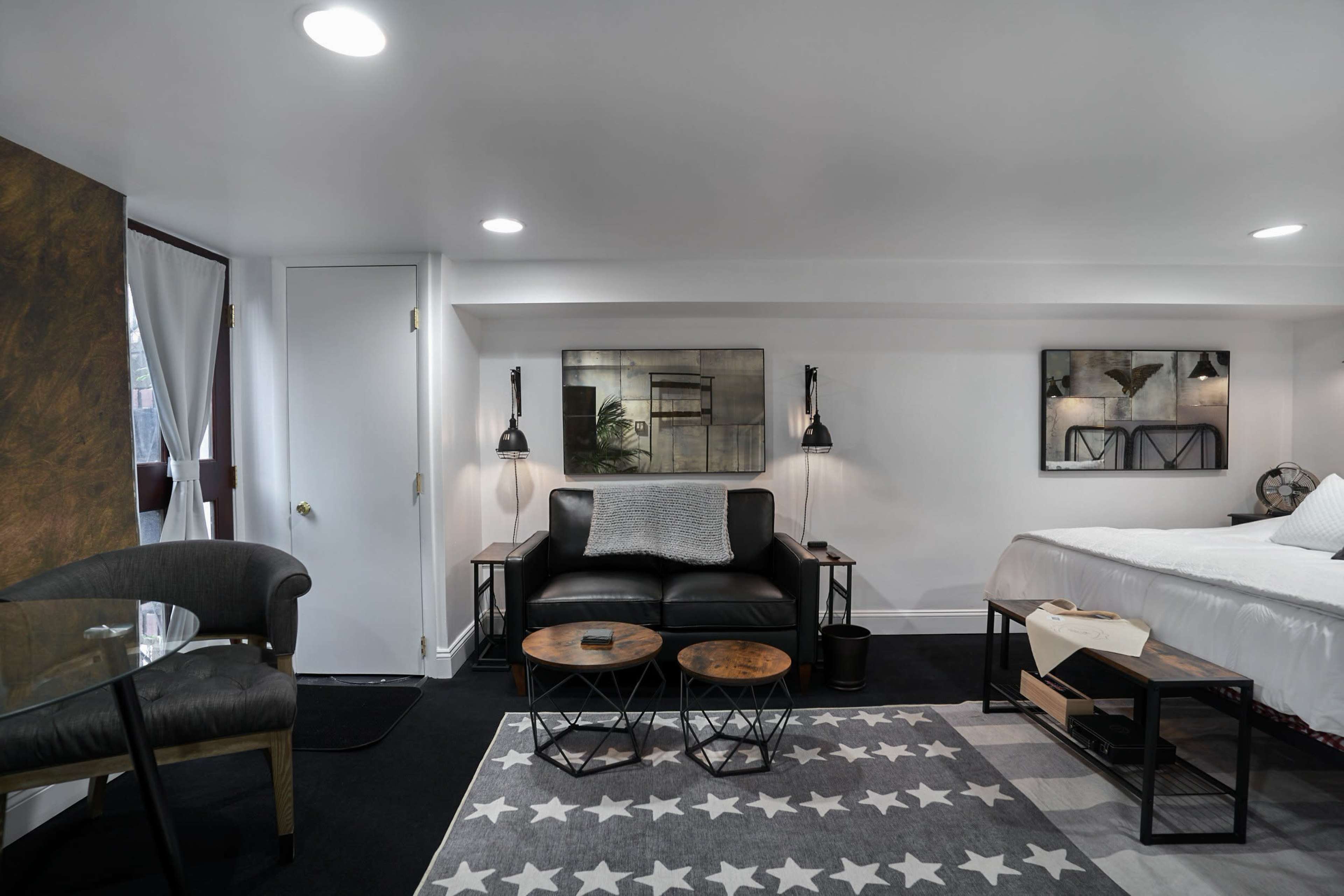 The image shows a modern hotel room featuring a black leather couch, a small round table, and a bed, with contemporary wall art and lamps providing illumination.