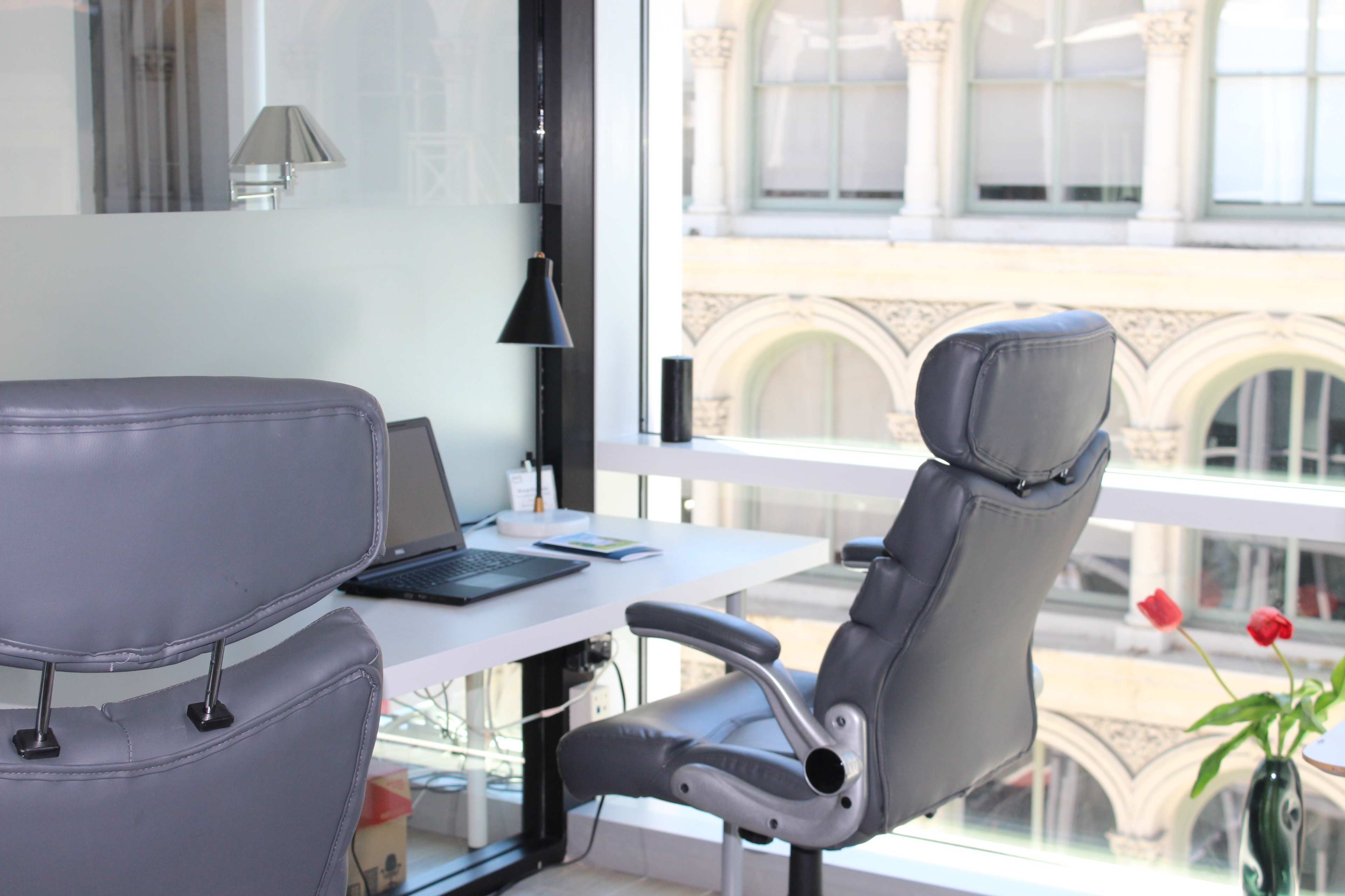 A modern office space features a desk with a laptop and a potted plant, overlooking a cityscape through large windows.