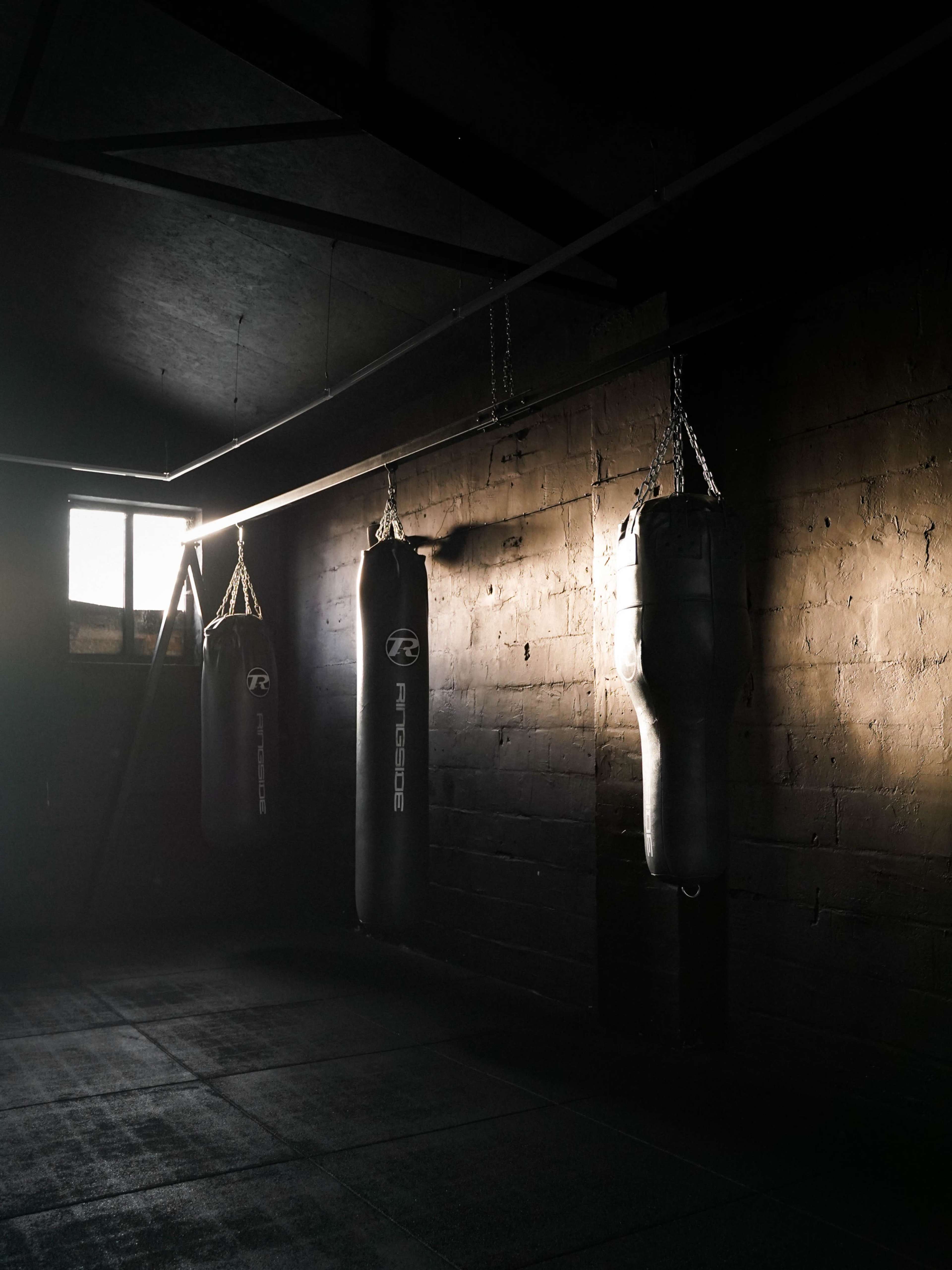 The image shows three hanging punching bags in a dimly lit gym with a window casting light on the wall.