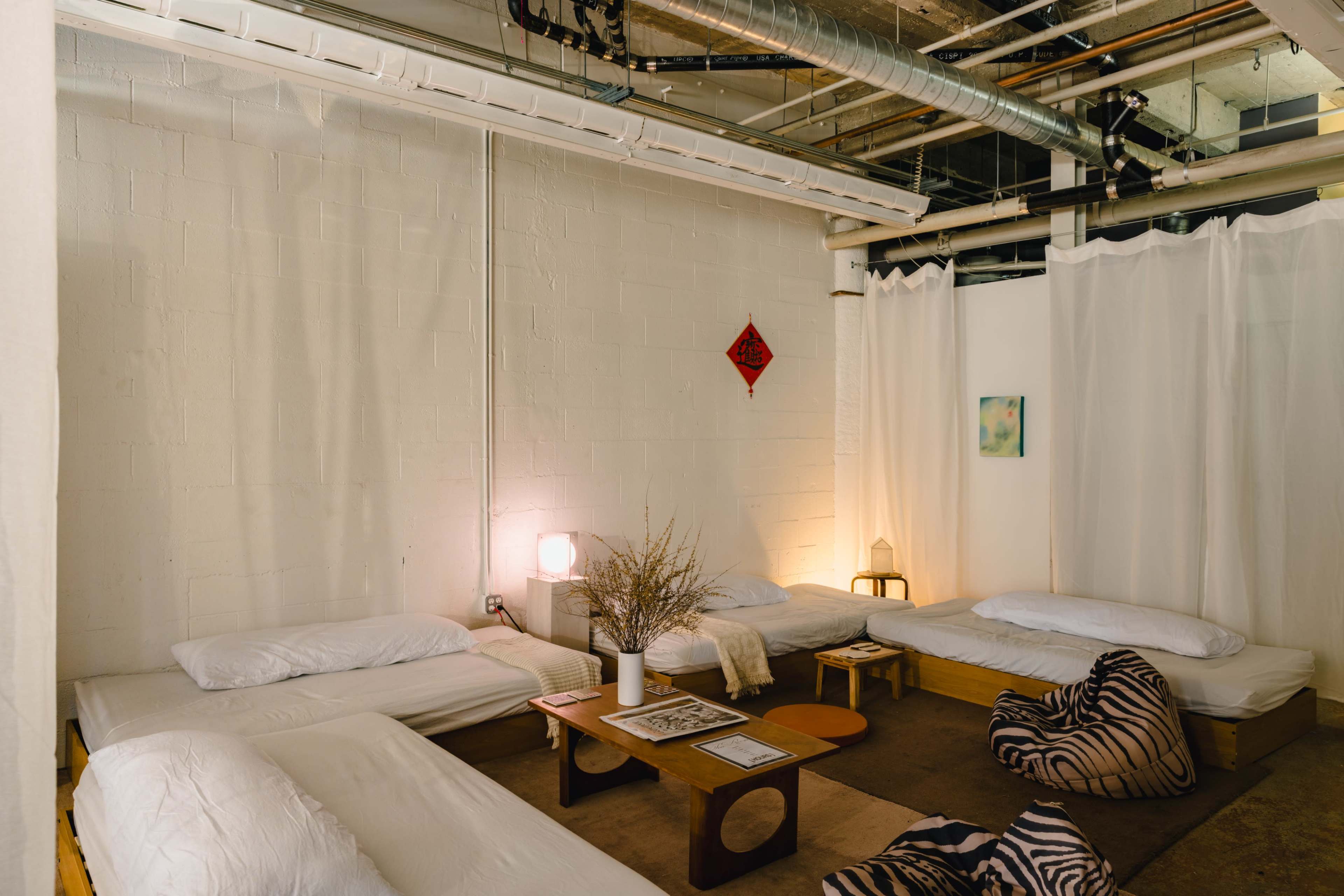 The image shows a minimalist indoor space with several low beds, a small table, and ambient lighting, separated by white curtains.