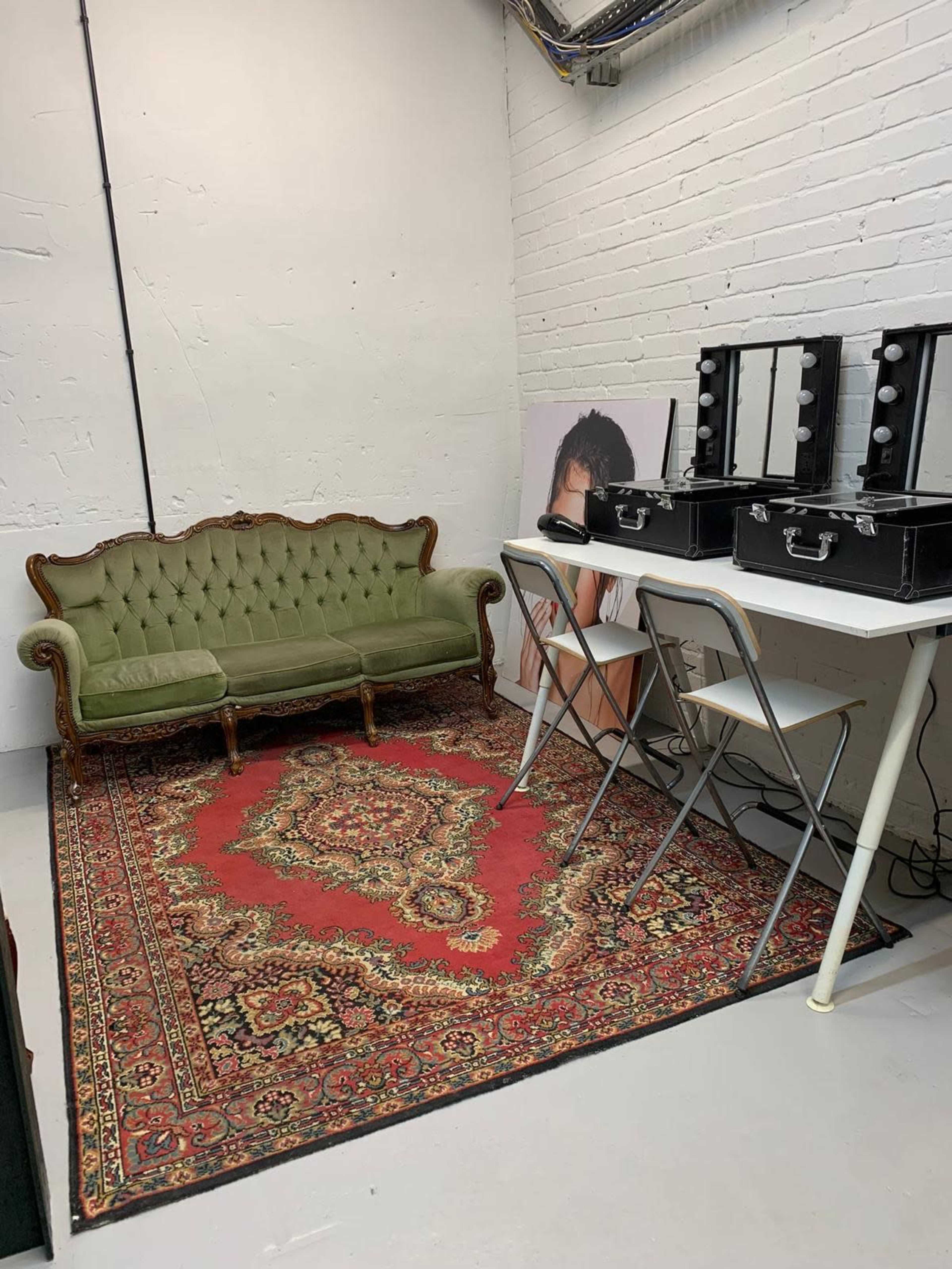 The image shows a small room with a green vintage sofa, a patterned rug, a white table with folding chairs, and two black vanity cases.