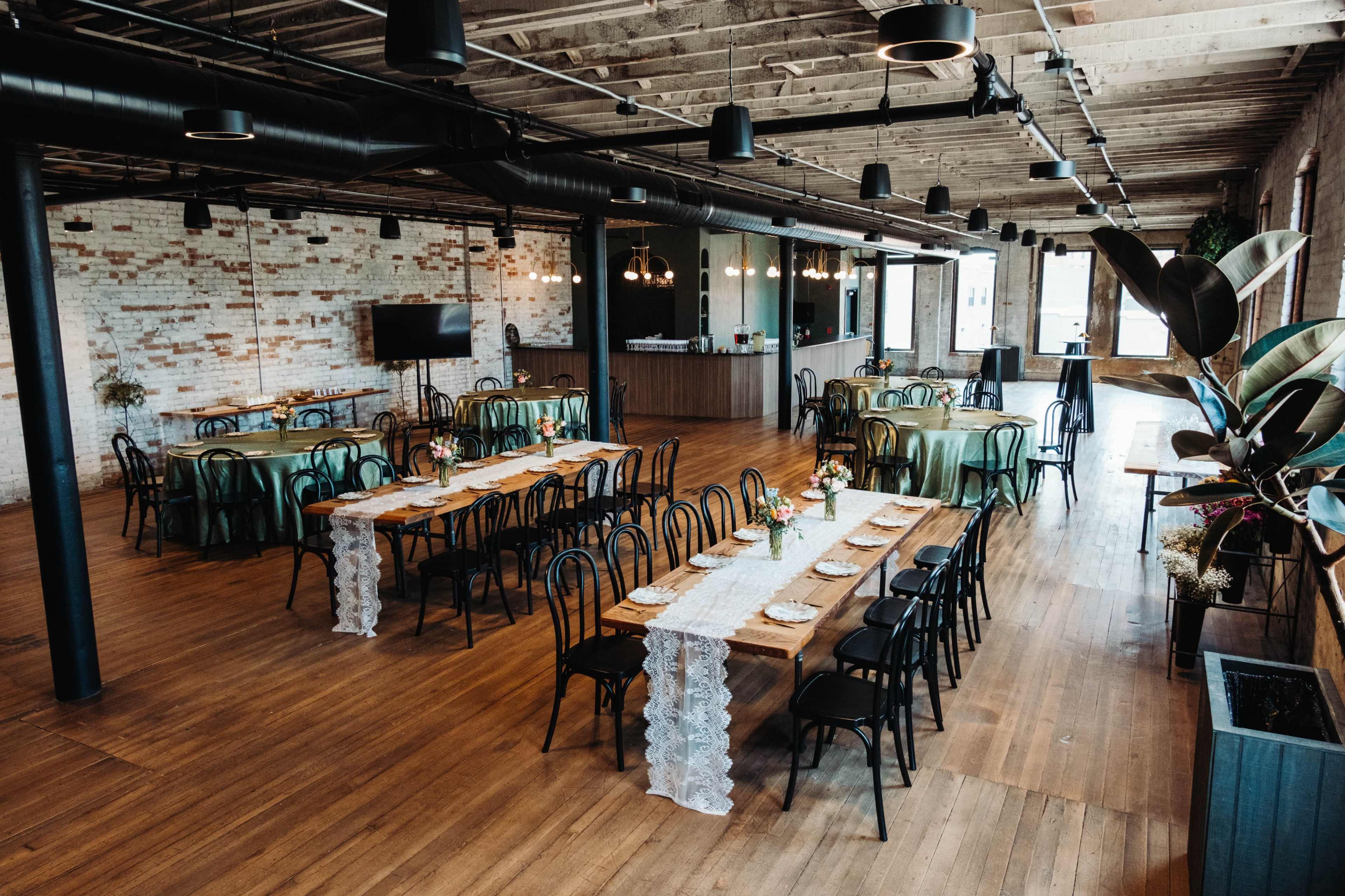 The image shows a spacious venue with wooden floors and tables set for a gathering, featuring green tablecloths and floral centerpieces.