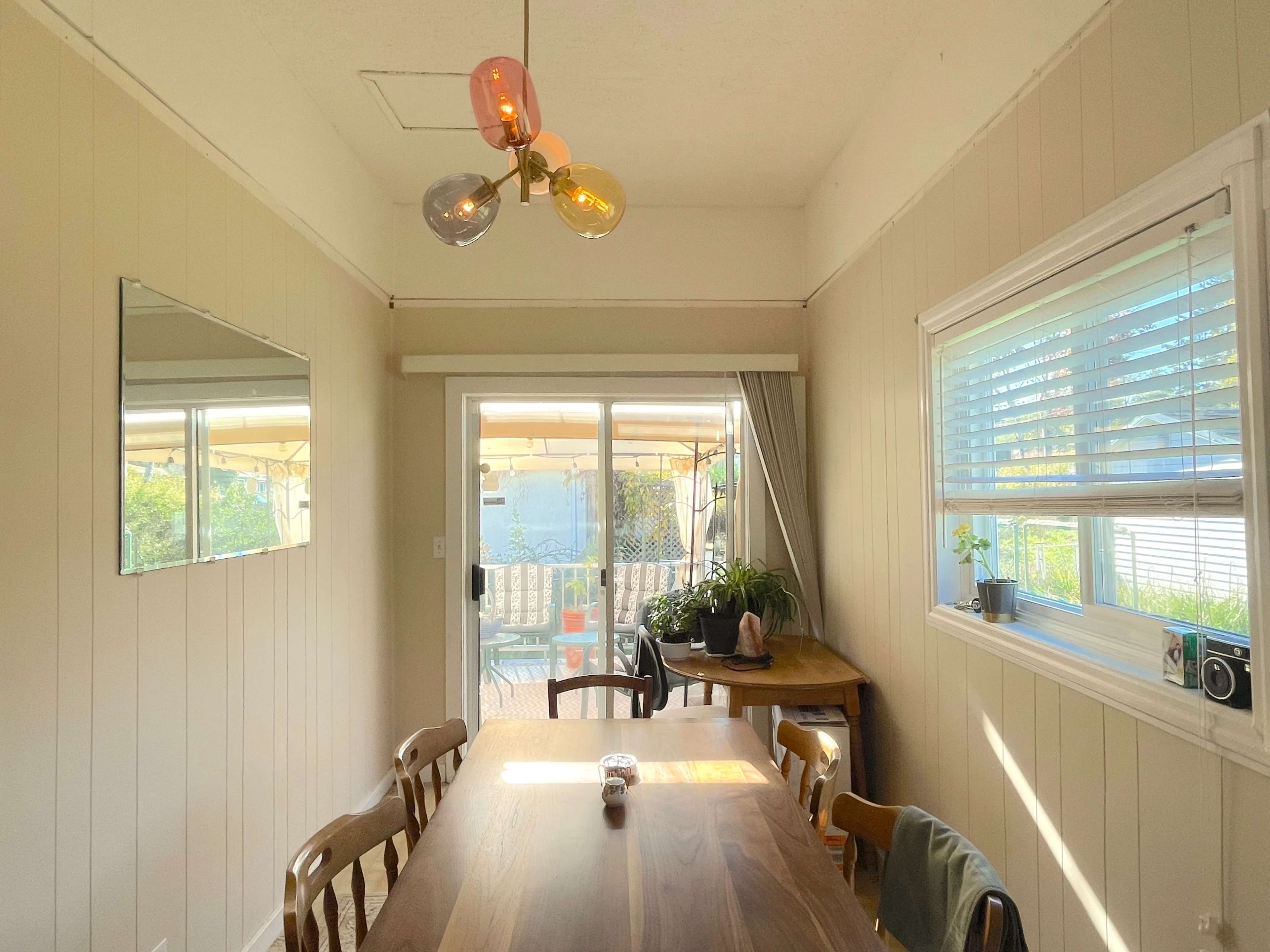 A simple dining area features a wooden table, four chairs, a large mirror on the wall, and glass light fixtures above.