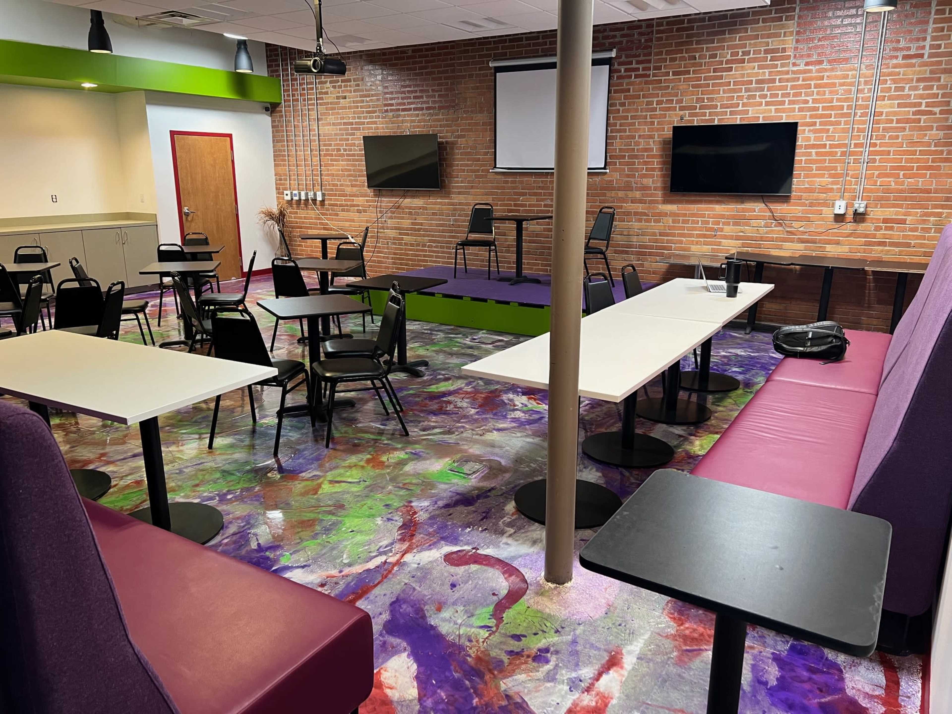 A room with a colorful floor, featuring tables and chairs arranged for seating, a small stage area, and two wall-mounted televisions.