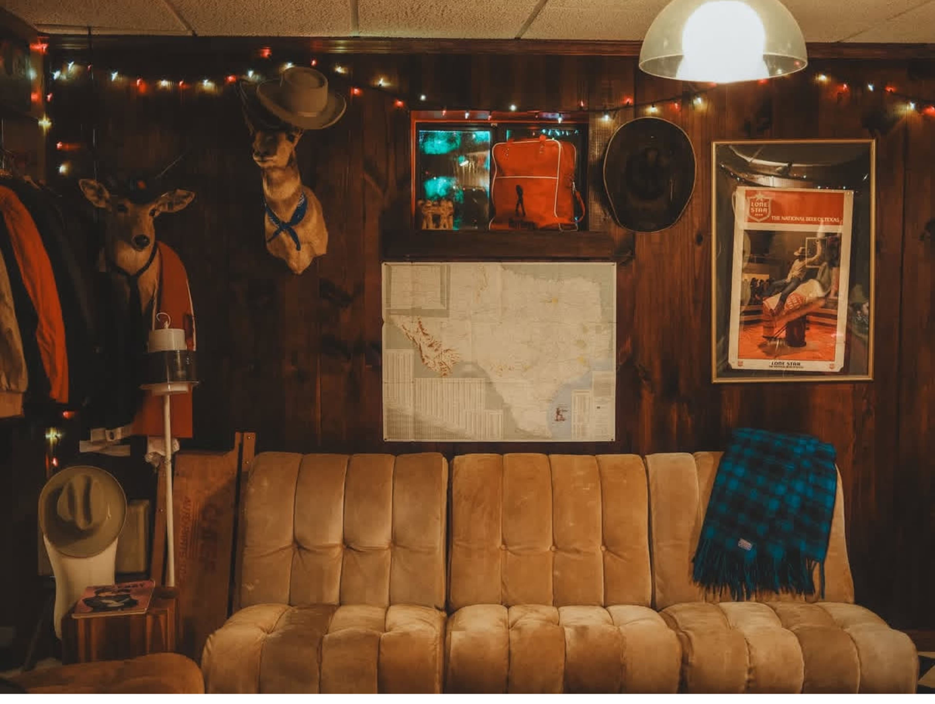 The image shows a cozy, rustic living space featuring a beige couch, a map on the wall, decorative lighting, and various mounted items, including deer heads and hats.