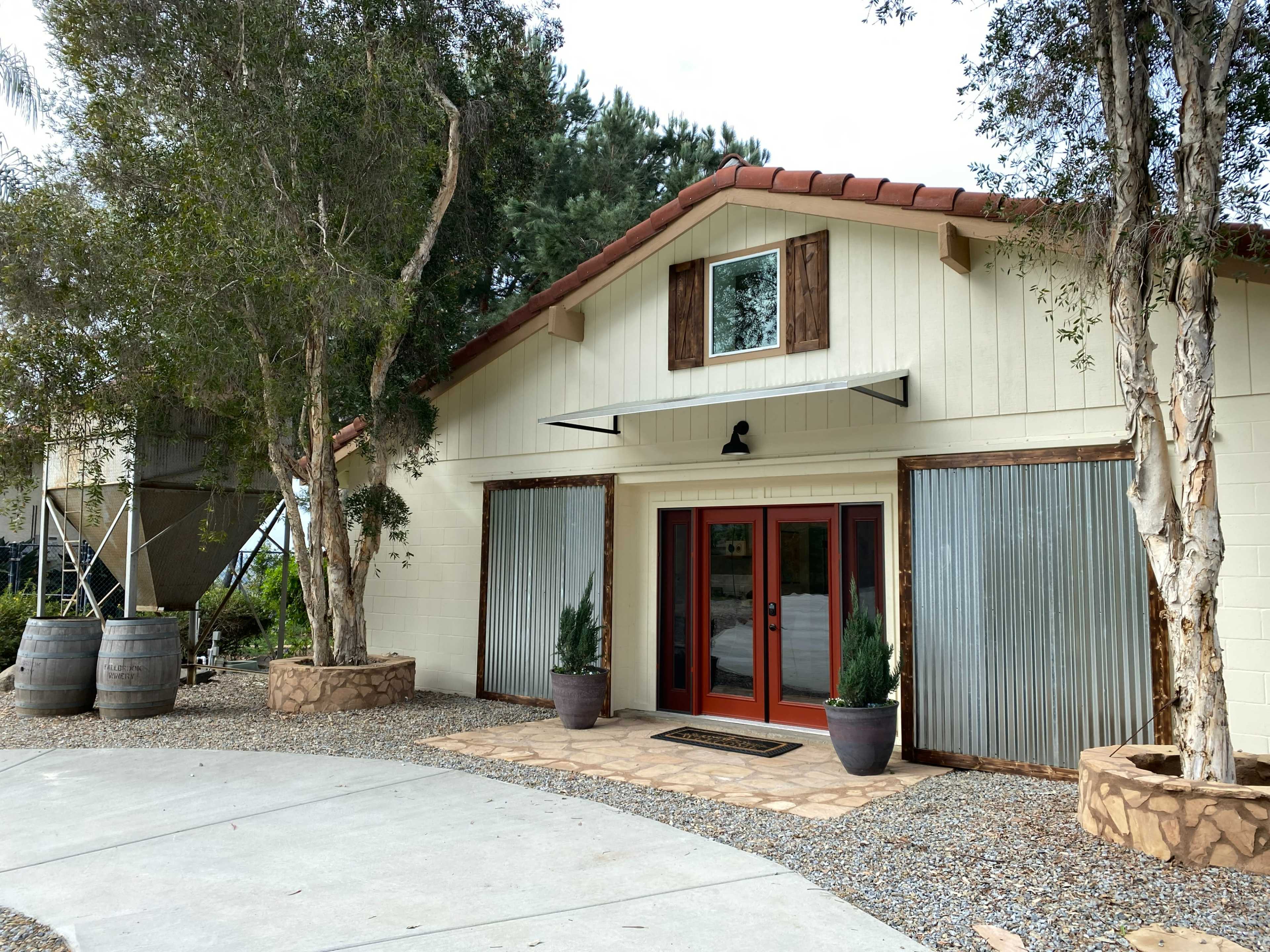 A modern building with a white exterior, large red double doors, and decorative metal panels on either side, surrounded by landscaped greenery and stone planters.