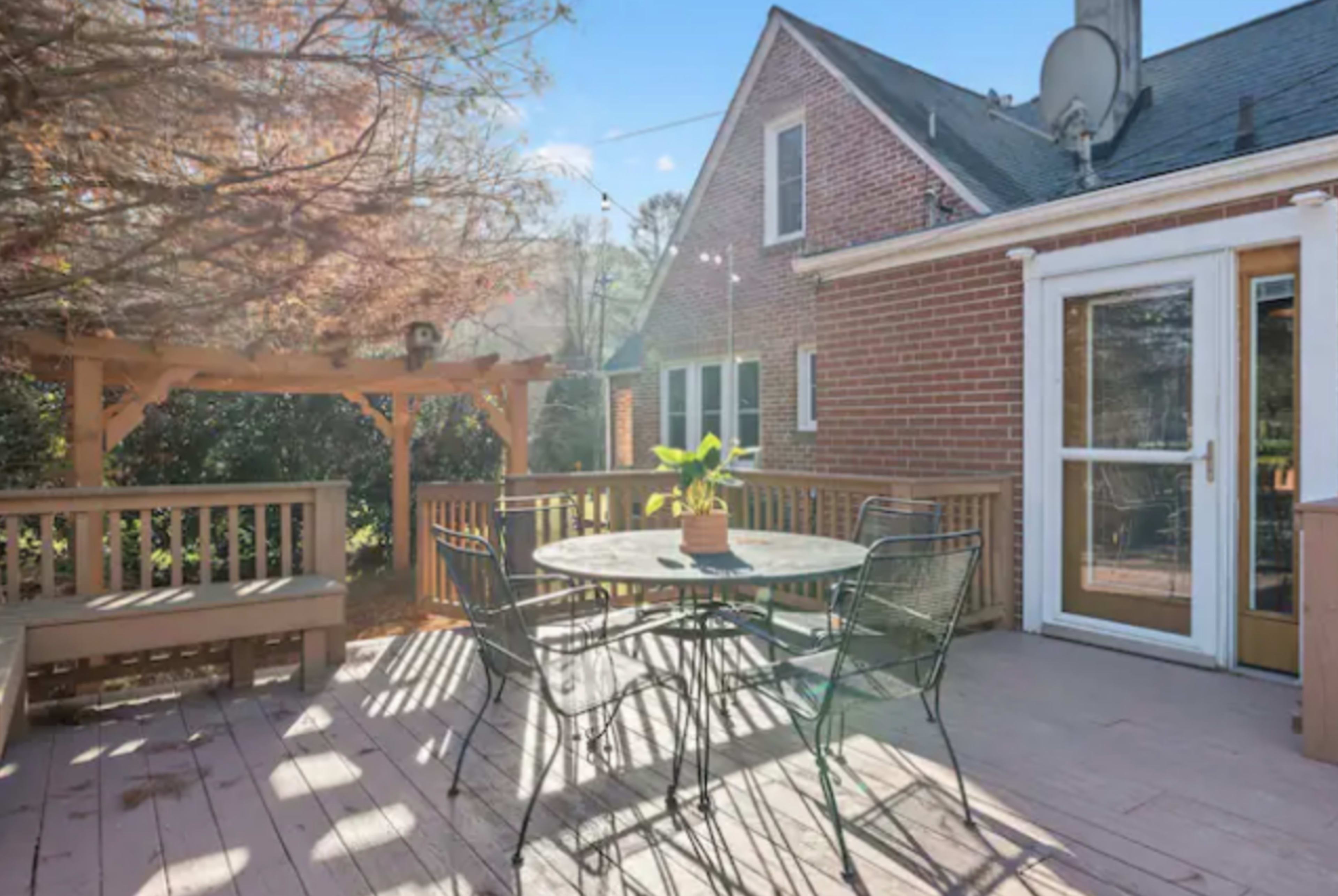 A wooden deck with a circular table and chairs, surrounded by a brick house and a pergola.