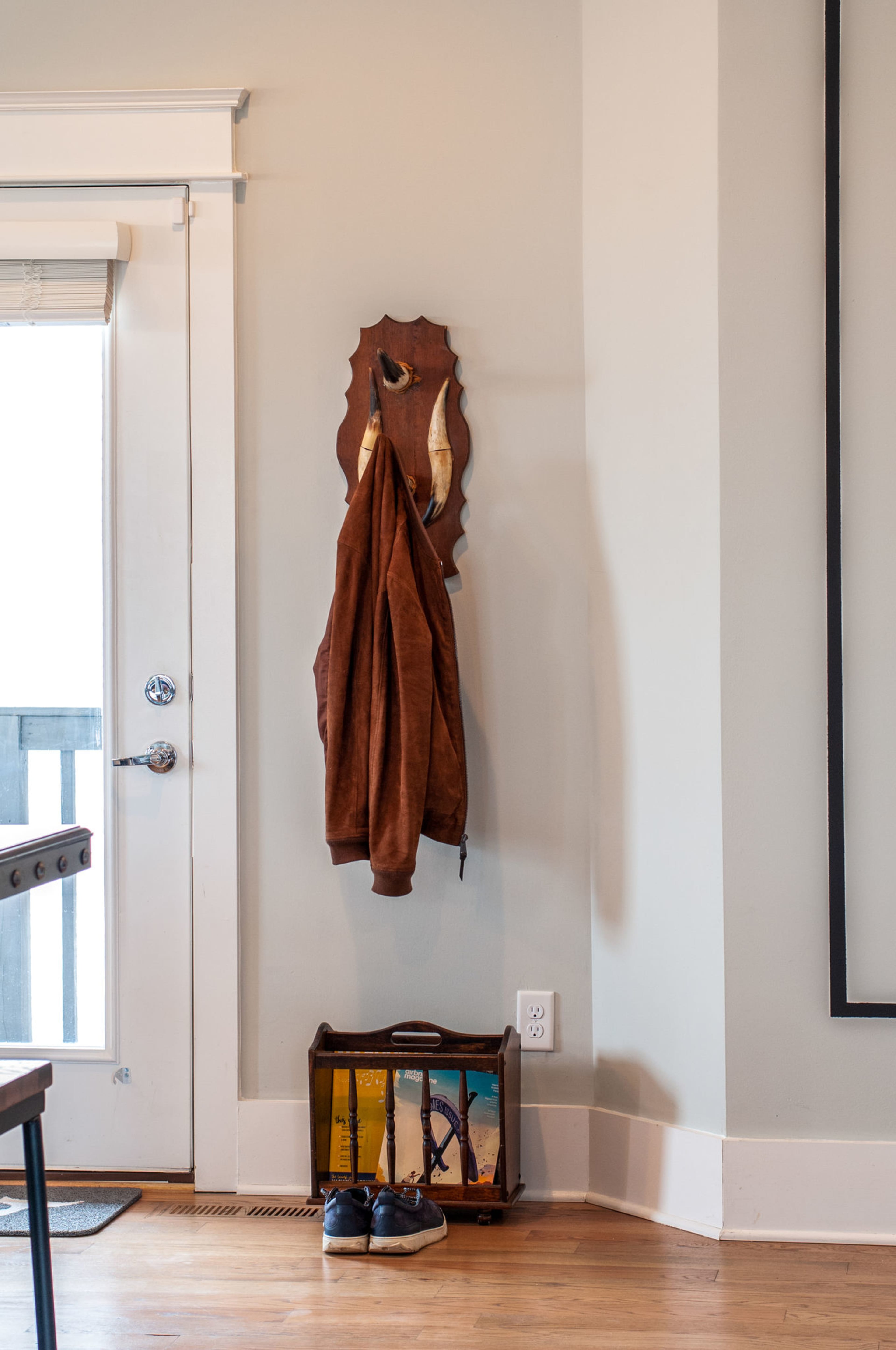 A brown jacket is hung on a wooden wall hook beside a door, with a small crate of books and a pair of shoes placed below it.