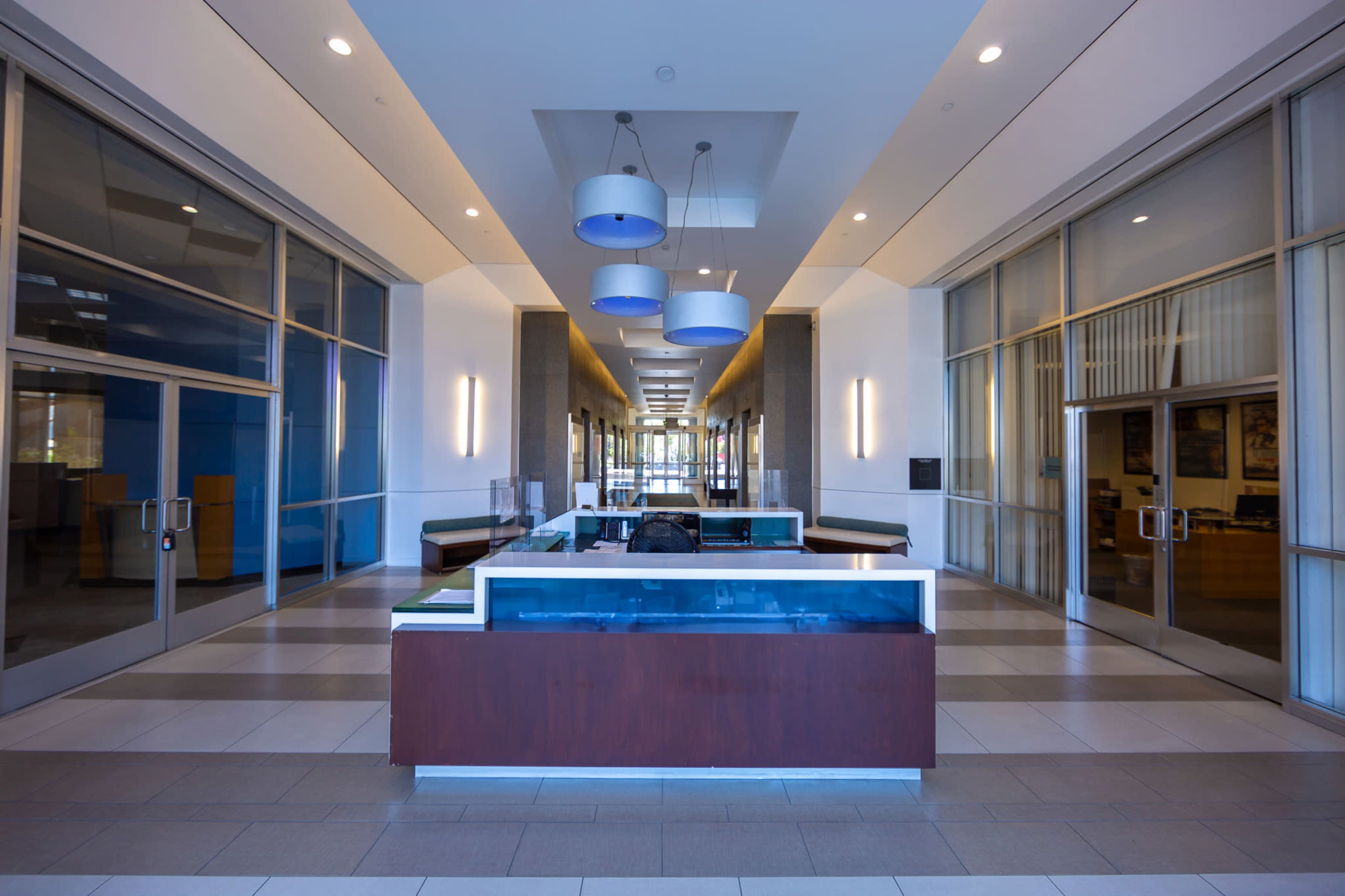 The image shows a modern office lobby featuring a reception desk in the center, flanked by large windows and overhead lighting.