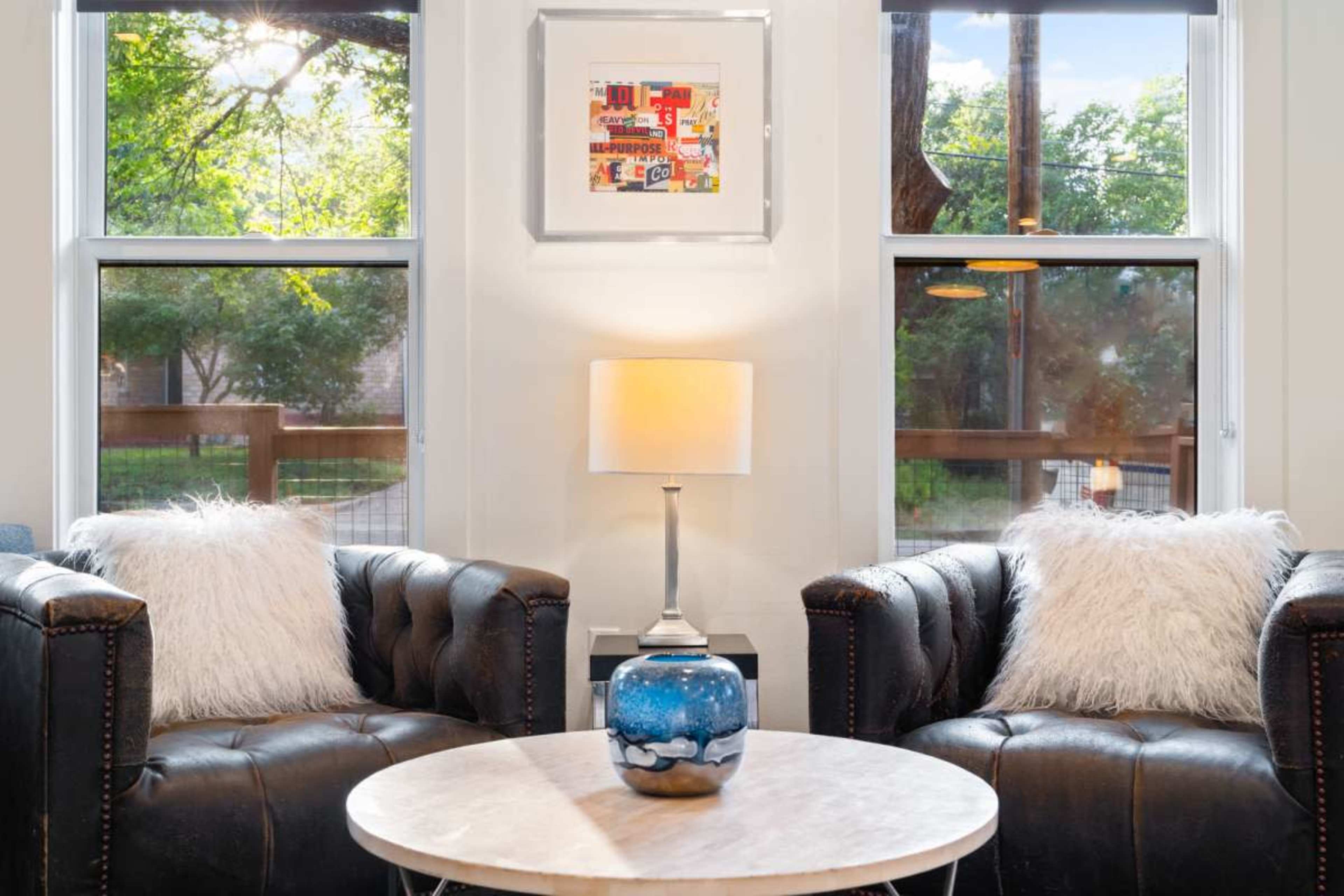 A cozy living room features two dark leather armchairs flanking a round marble table, with a lamp on the table and colorful artwork displayed on the wall above.