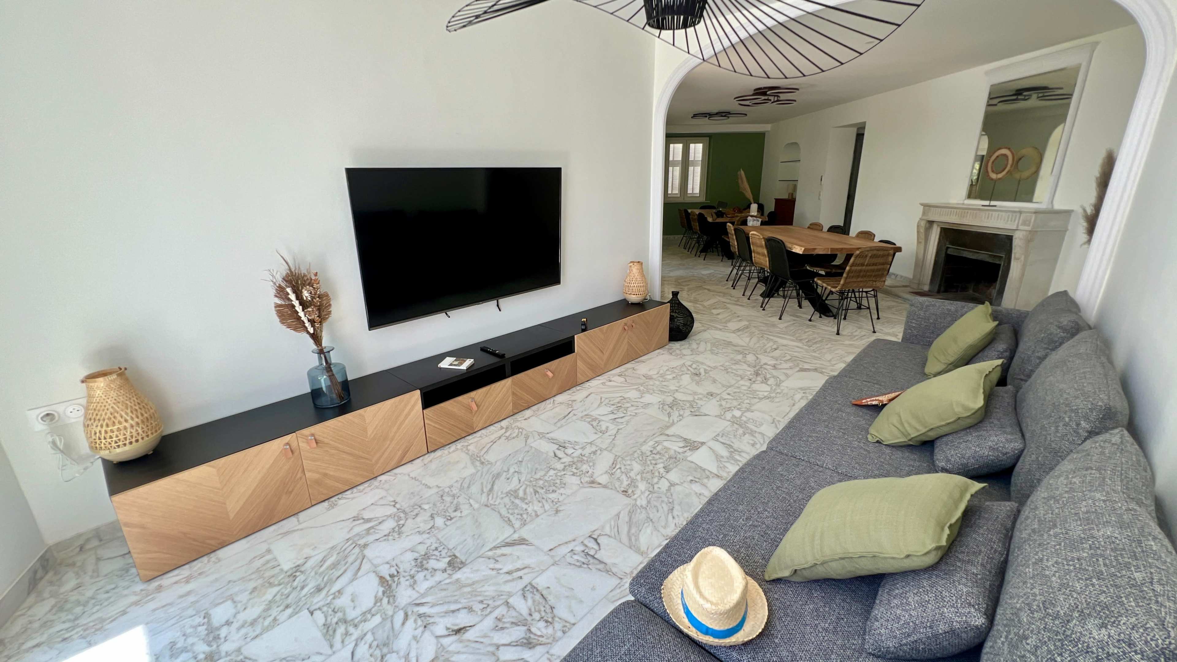 The image shows a modern living room with a gray sofa, a wooden TV stand, and a large flat-screen TV against a white wall, featuring a marble floor and open access to a dining area in the background.