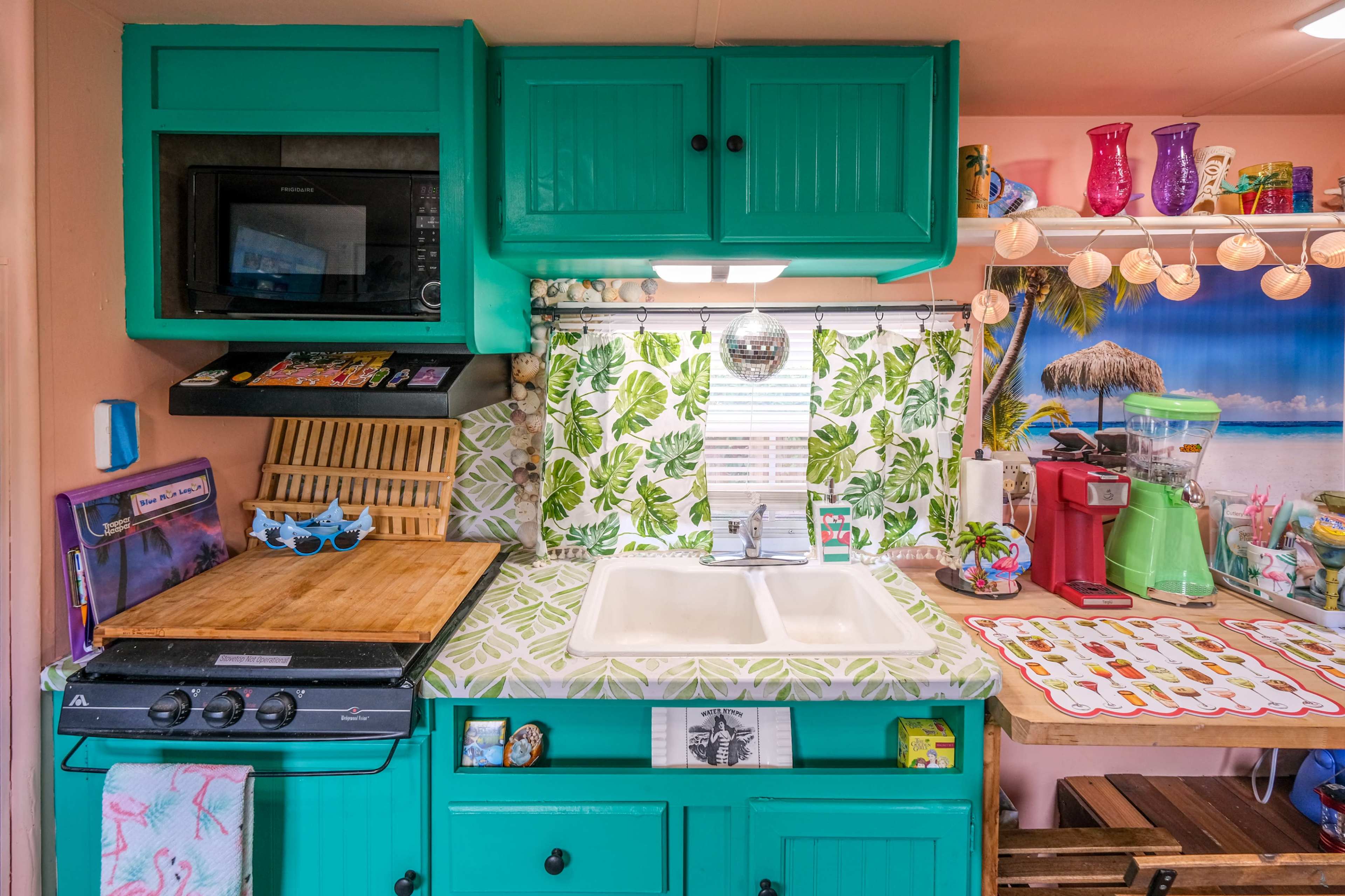 The image shows a compact kitchen with turquoise cabinets, a microwave, a stove, and various kitchen accessories amid a tropical-themed decor.
