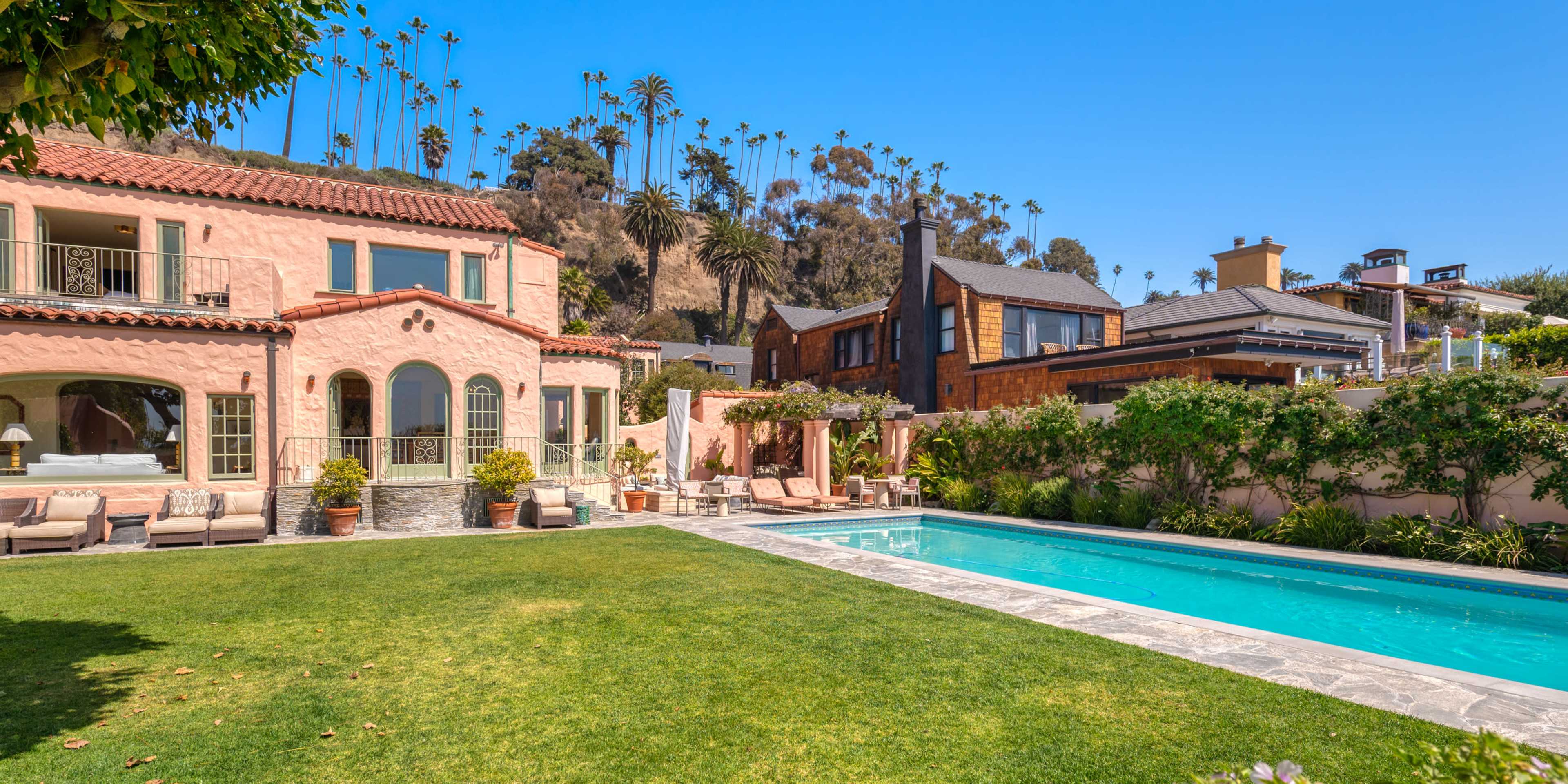A landscaped backyard features a swimming pool surrounded by a stone patio, with two large houses and palm trees in the background.