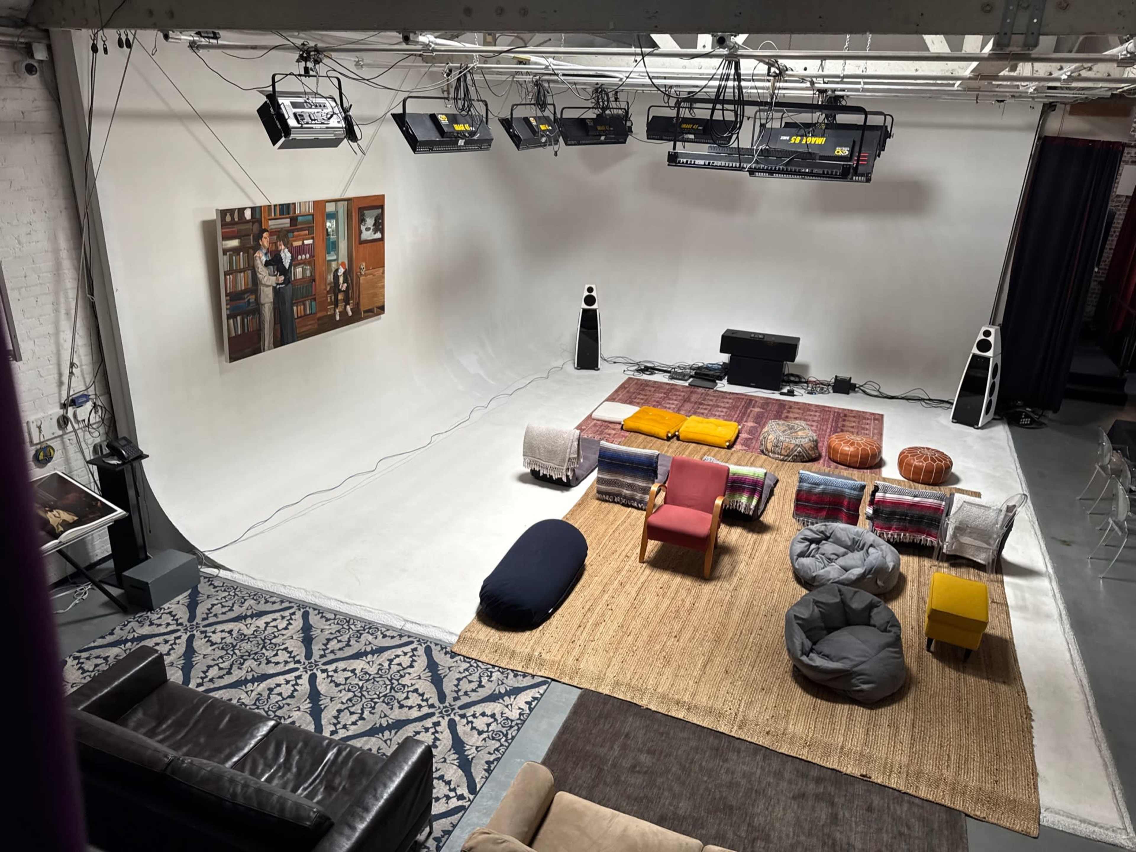 The image shows a spacious studio setup with various seating options, including plush chairs and ottomans, against a plain backdrop and equipped with overhead lighting.