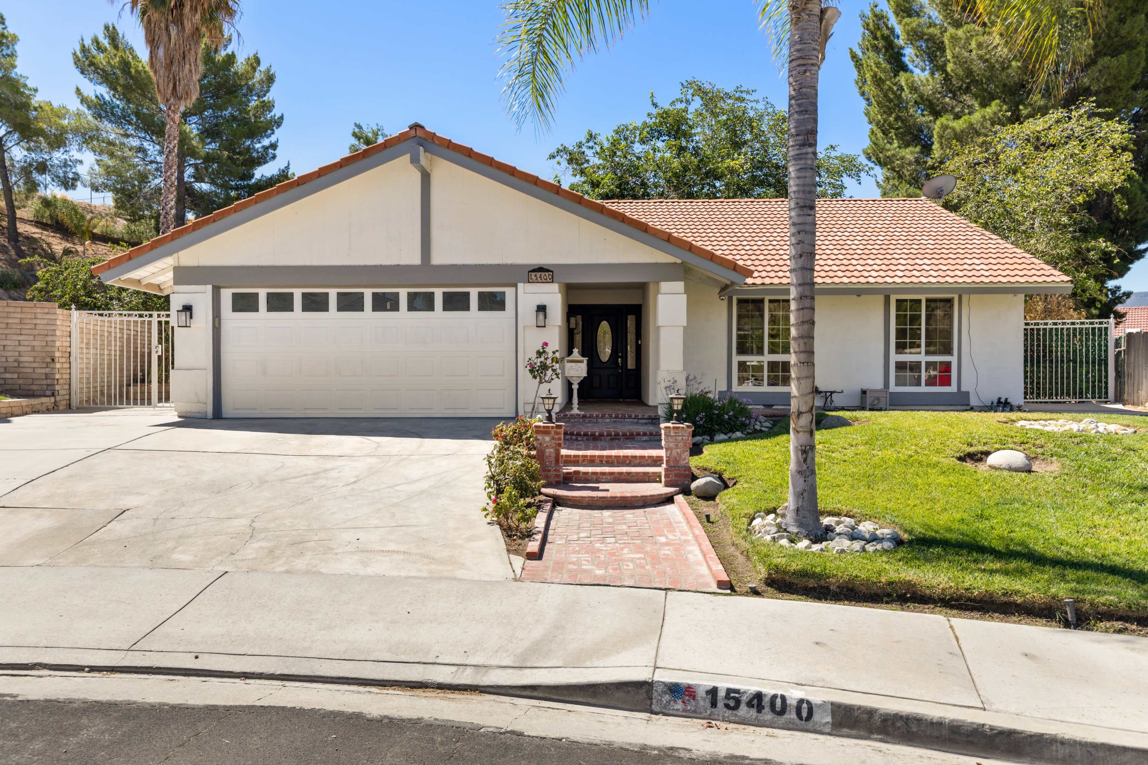 The image shows a single-story house with a brick pathway leading to the front door, a manicured lawn, and a two-car garage.