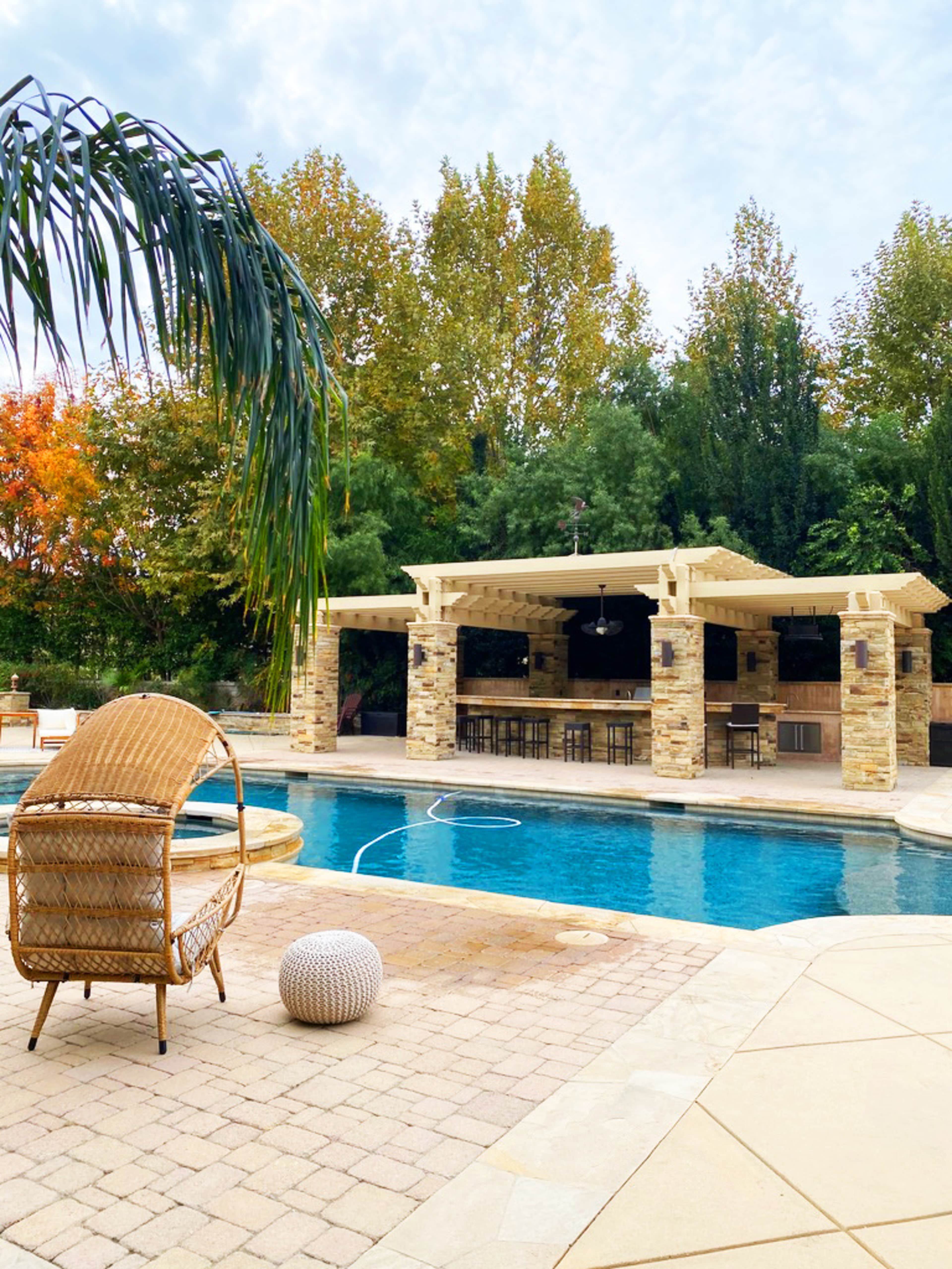A stone patio surrounds a swimming pool, with a covered outdoor kitchen and seating area visible in the background amid trees displaying autumn colors.