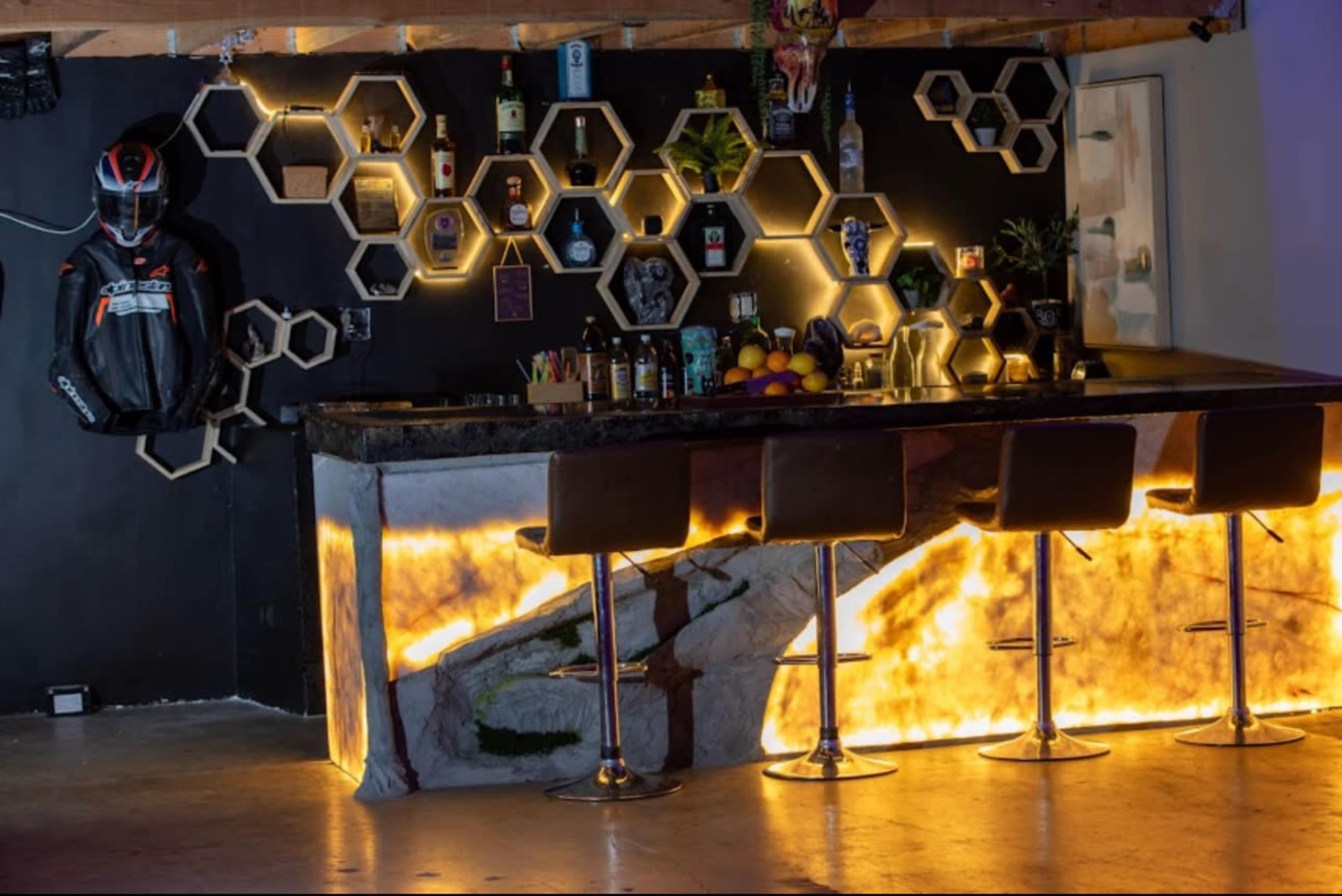 The image shows a modern bar setup featuring a hexagonal shelf display with various bottles and a backlit counter made of textured material, accompanied by sleek bar stools.
