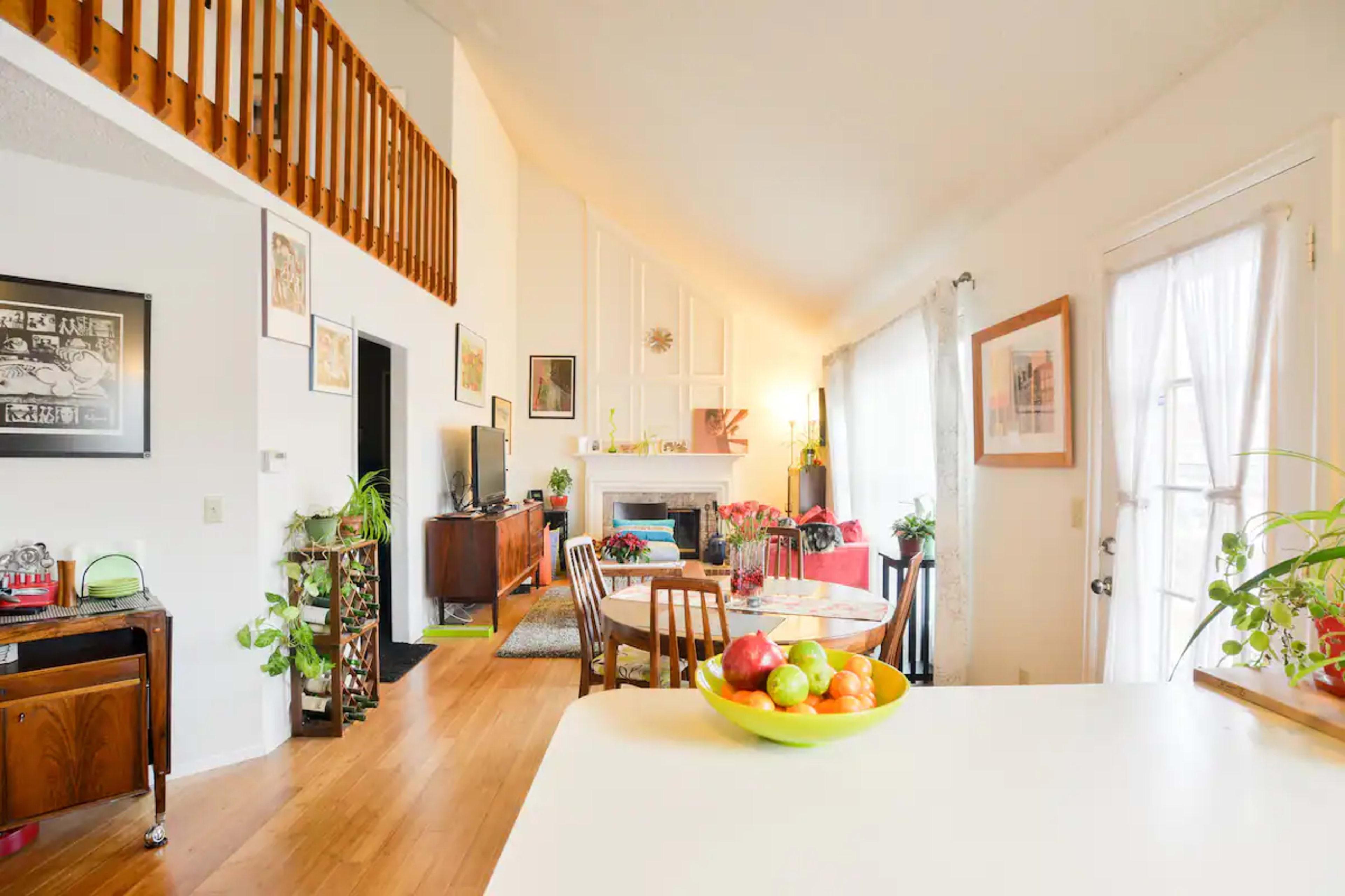 A bright, open-concept living space featuring a dining area, a kitchen counter with a bowl of fruit, and large windows letting in natural light.