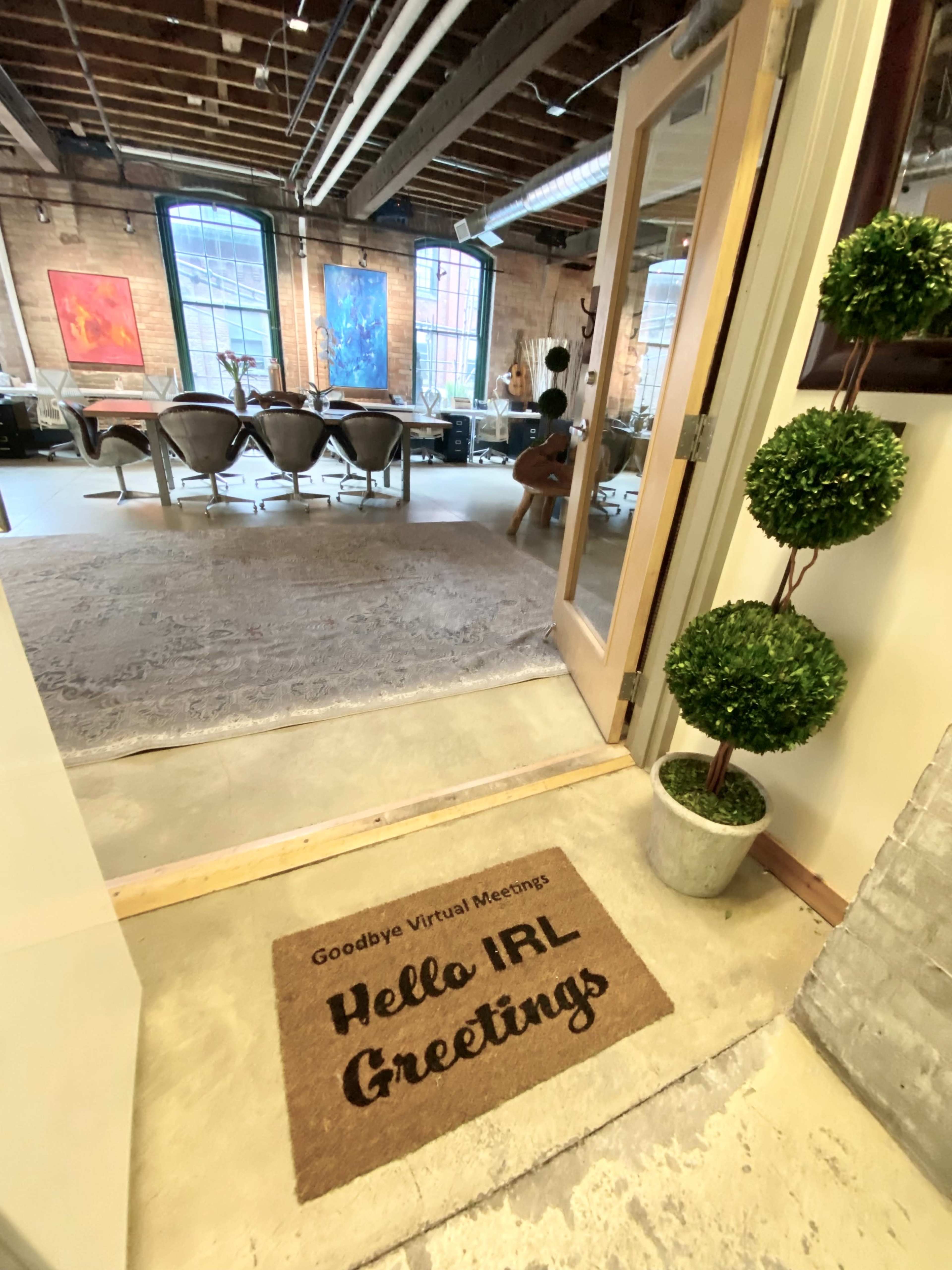 A doorway with a welcome mat stating "Hello IRL Greetings," leading into a spacious room with modern furniture and artwork on the walls.