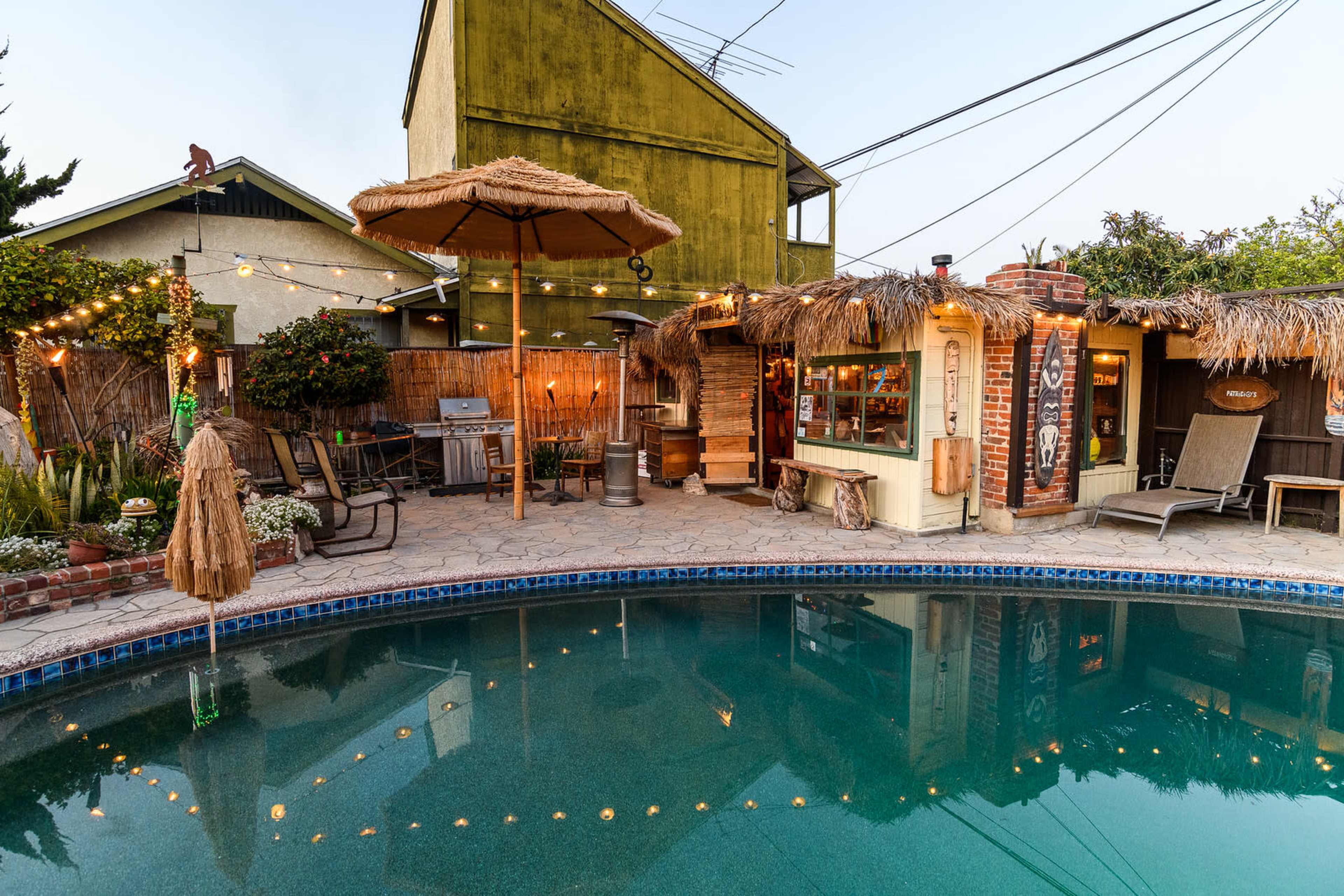 A backyard with a pool, a thatched umbrella, string lights, and a small outdoor bar area next to a house.
