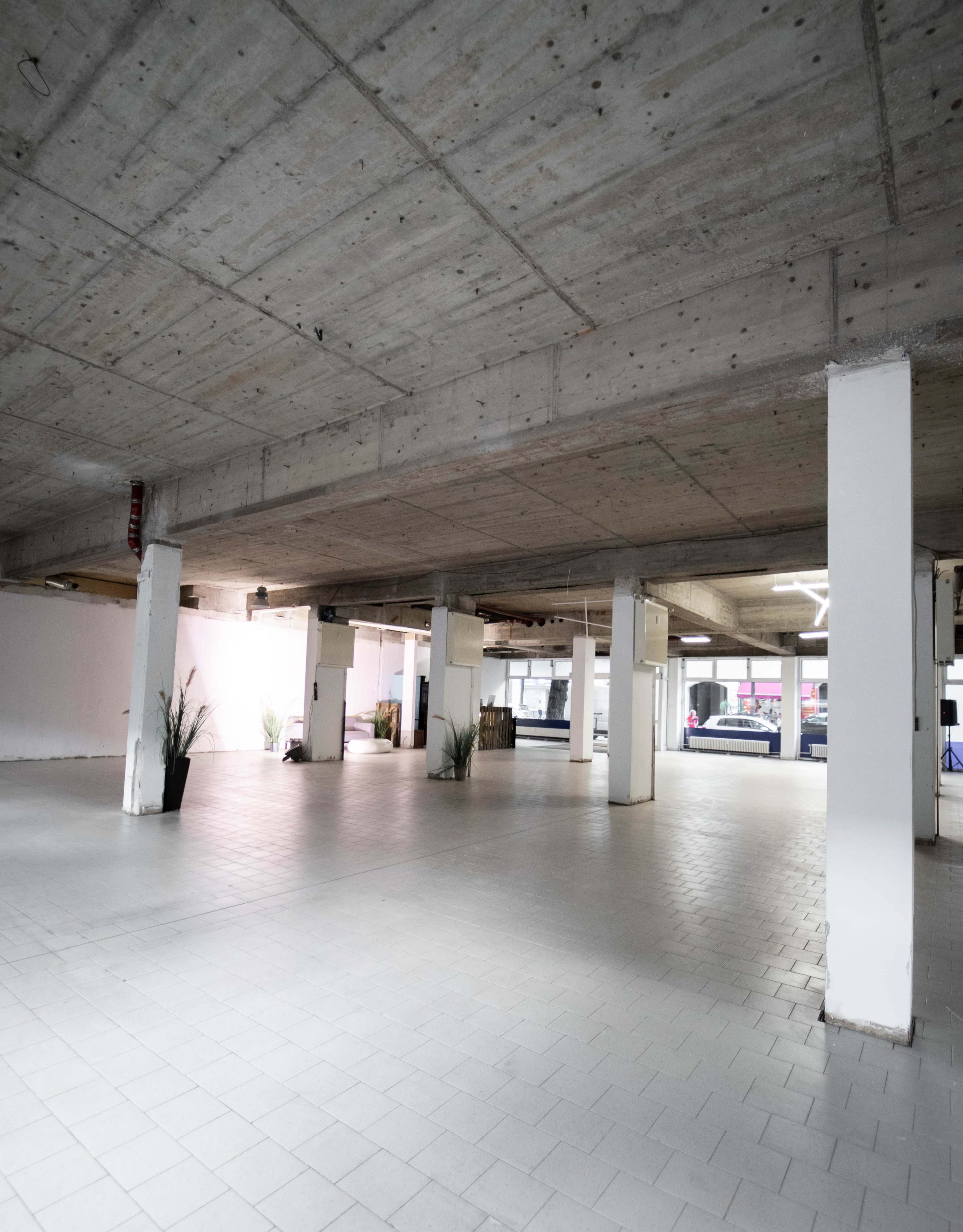 A large, empty indoor space with concrete walls and floor tiles, featuring several columns and minimal furnishings.