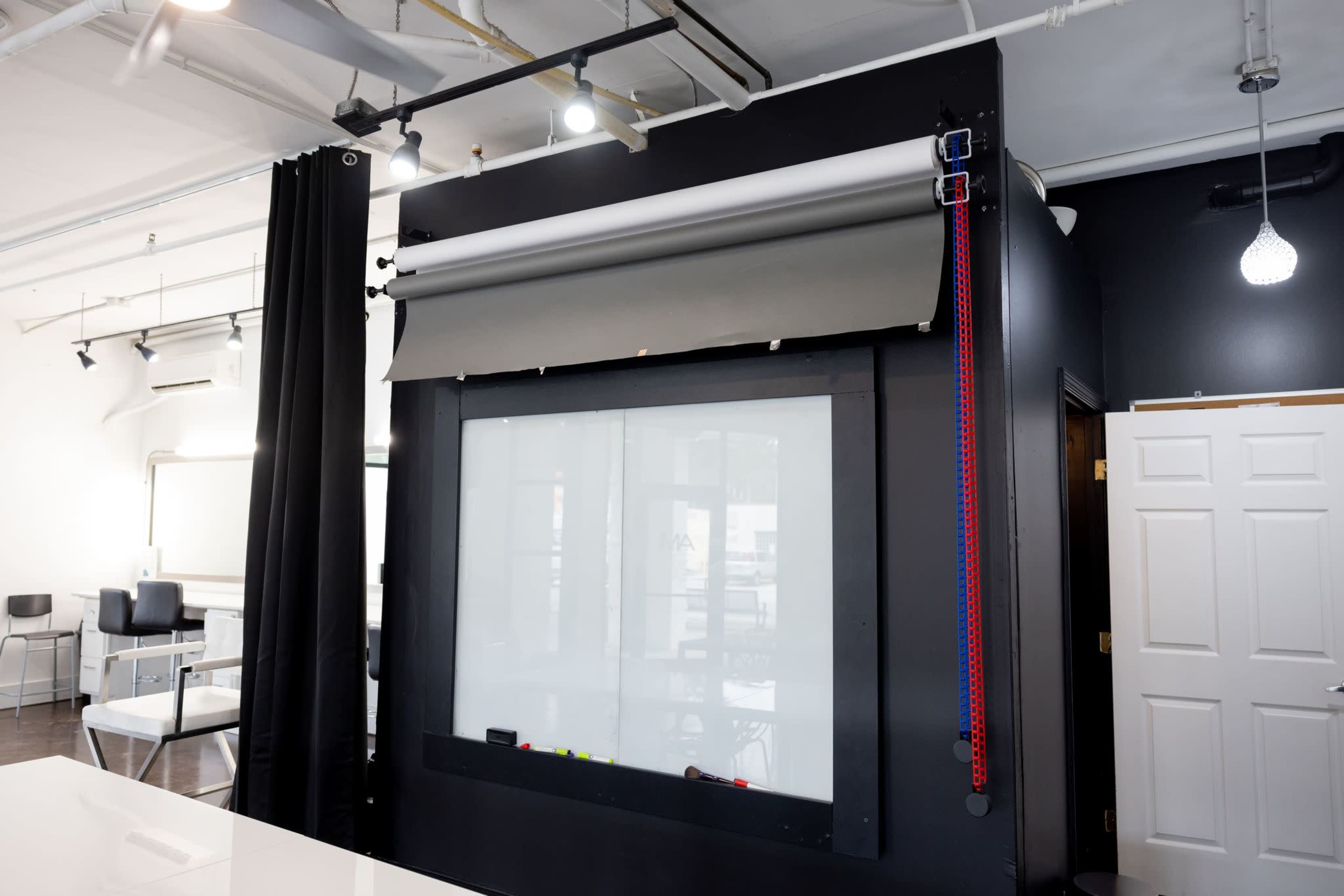 A black wall with a retractable projection screen, flanked by a curtain and a white door, inside a well-lit workspace.