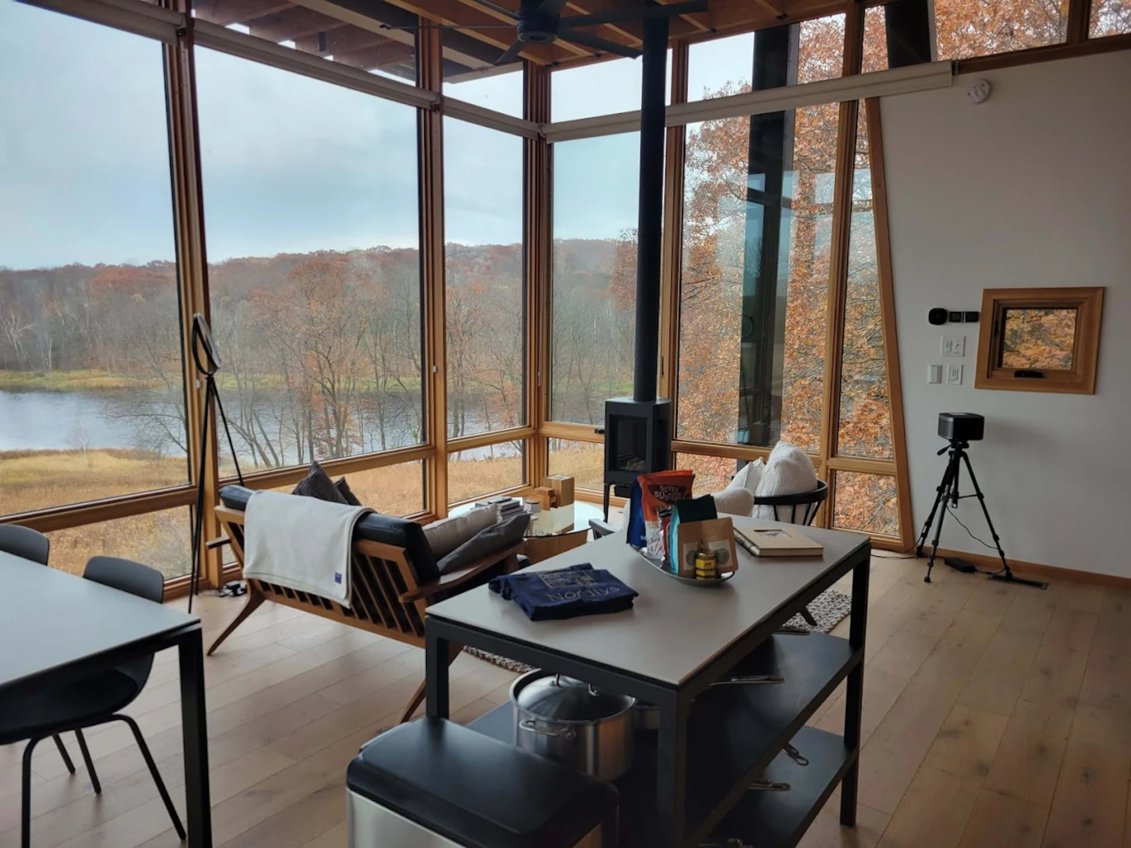 A modern living space features large windows overlooking a forested landscape with autumn foliage, a minimalist seating area, and a dining table.