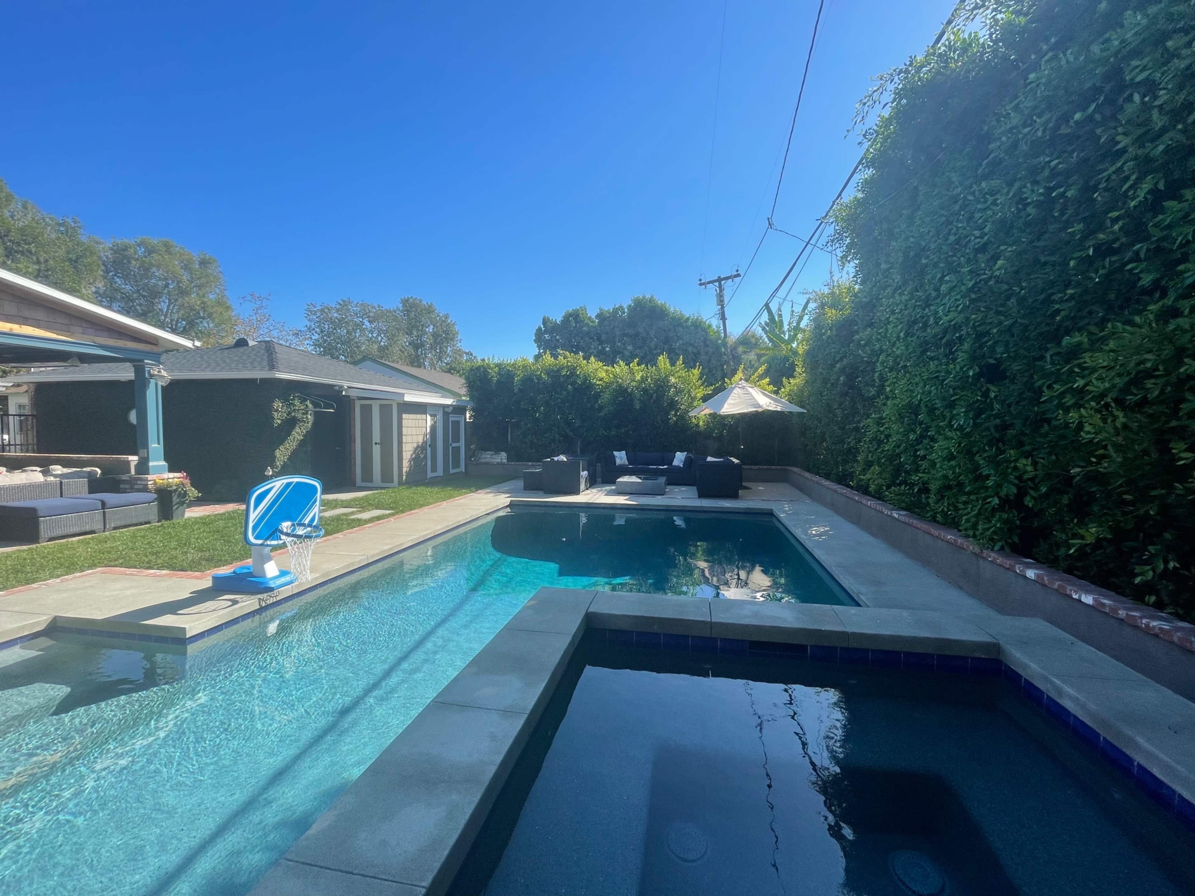 The image shows a backyard with a swimming pool, a hot tub, lounge seating, and lush greenery surrounding the area.