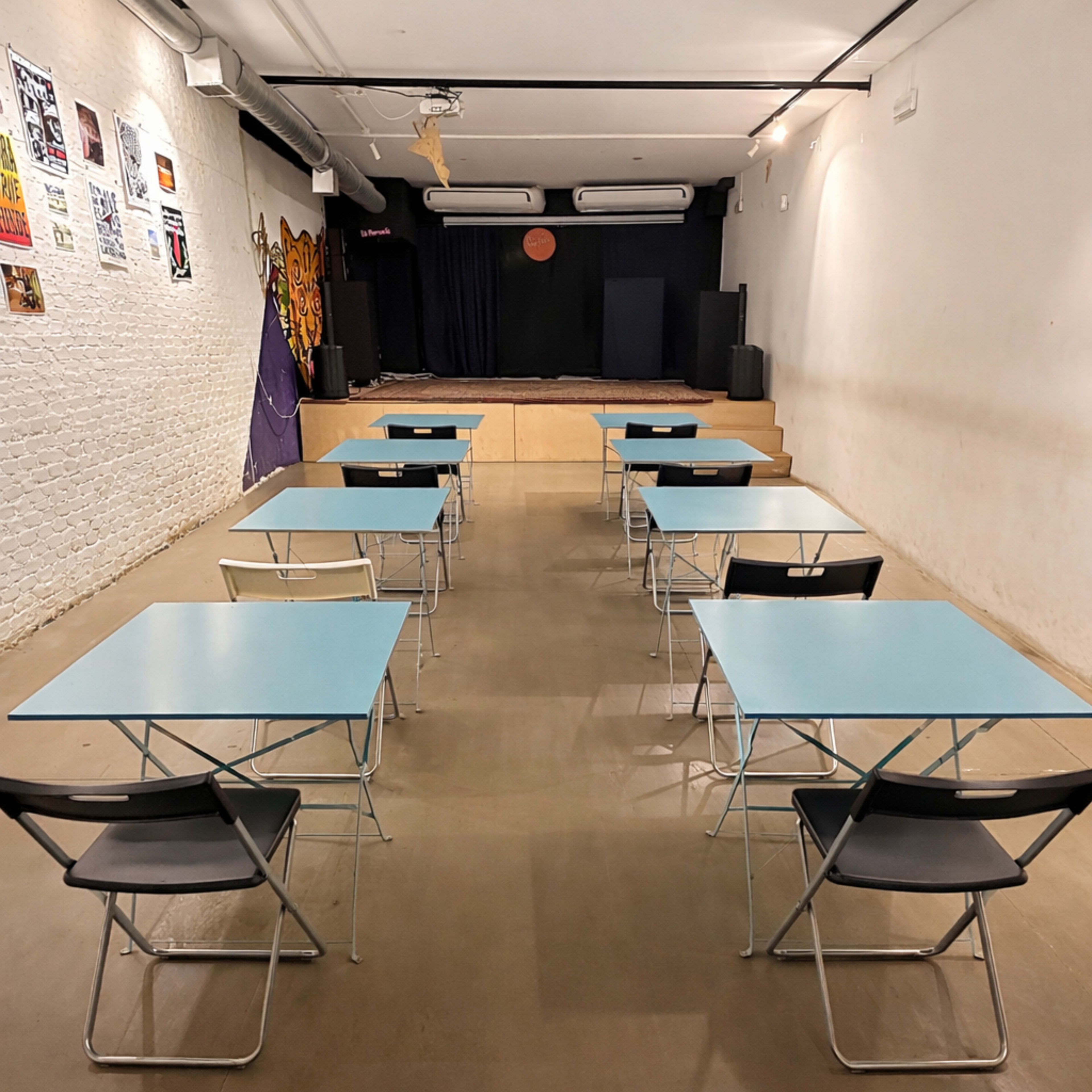 The image shows a vacant room set up with several blue folding tables and chairs facing a small stage area at the front.