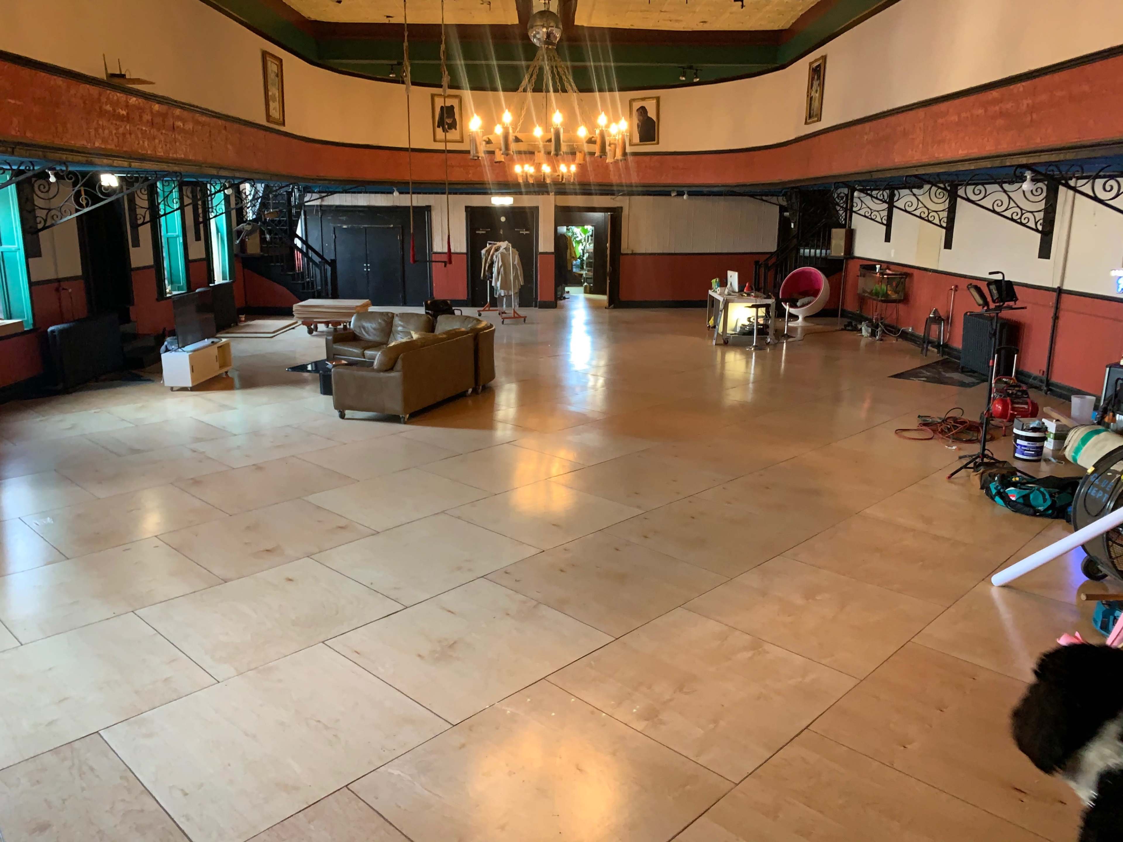 The image shows a spacious indoor area with wooden flooring, featuring a few pieces of furniture, including a brown couch and a table, under a chandelier.