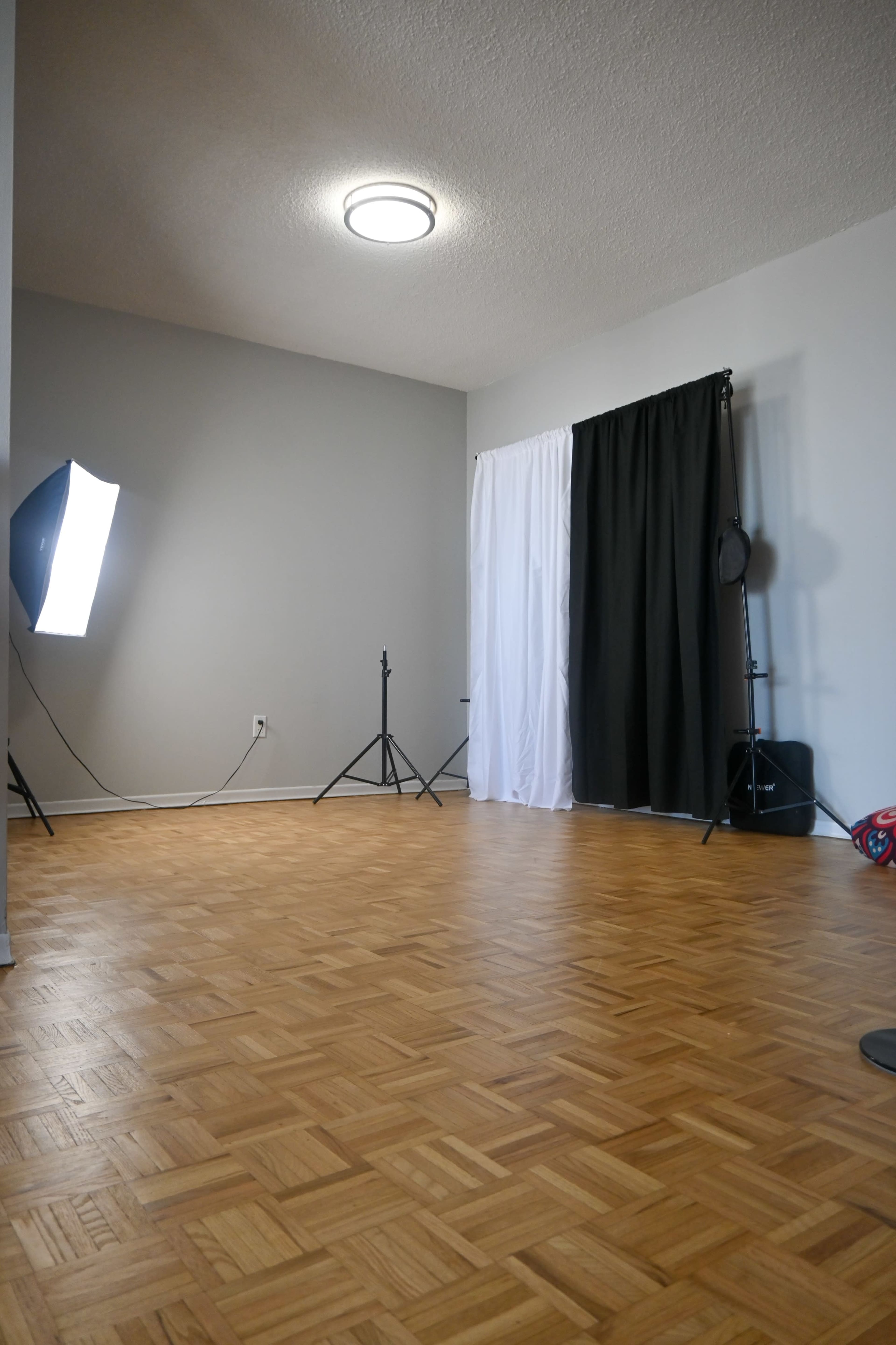The image shows an empty room with a wooden parquet floor, featuring a gray wall, a light fixture on the ceiling, and two fabric backdrops in black and white.