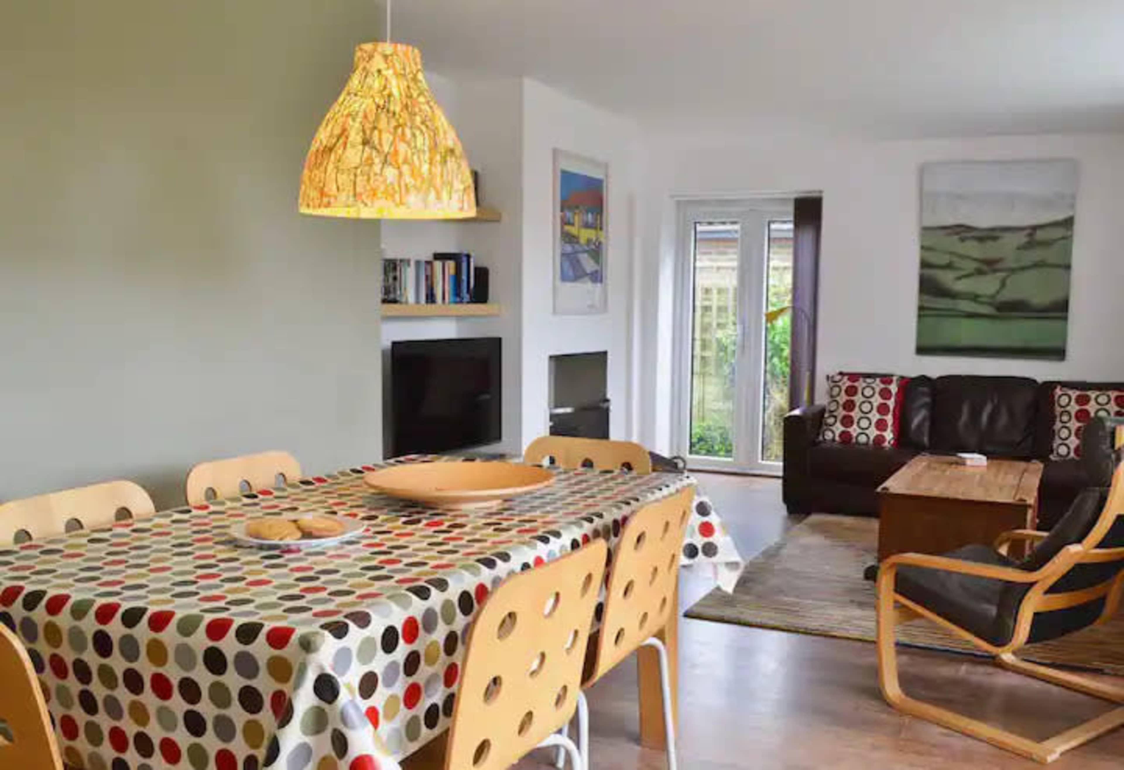 The image shows a modern dining area with a table set for meals, surrounded by chairs, leading into a cozy living space with a couch and a coffee table.