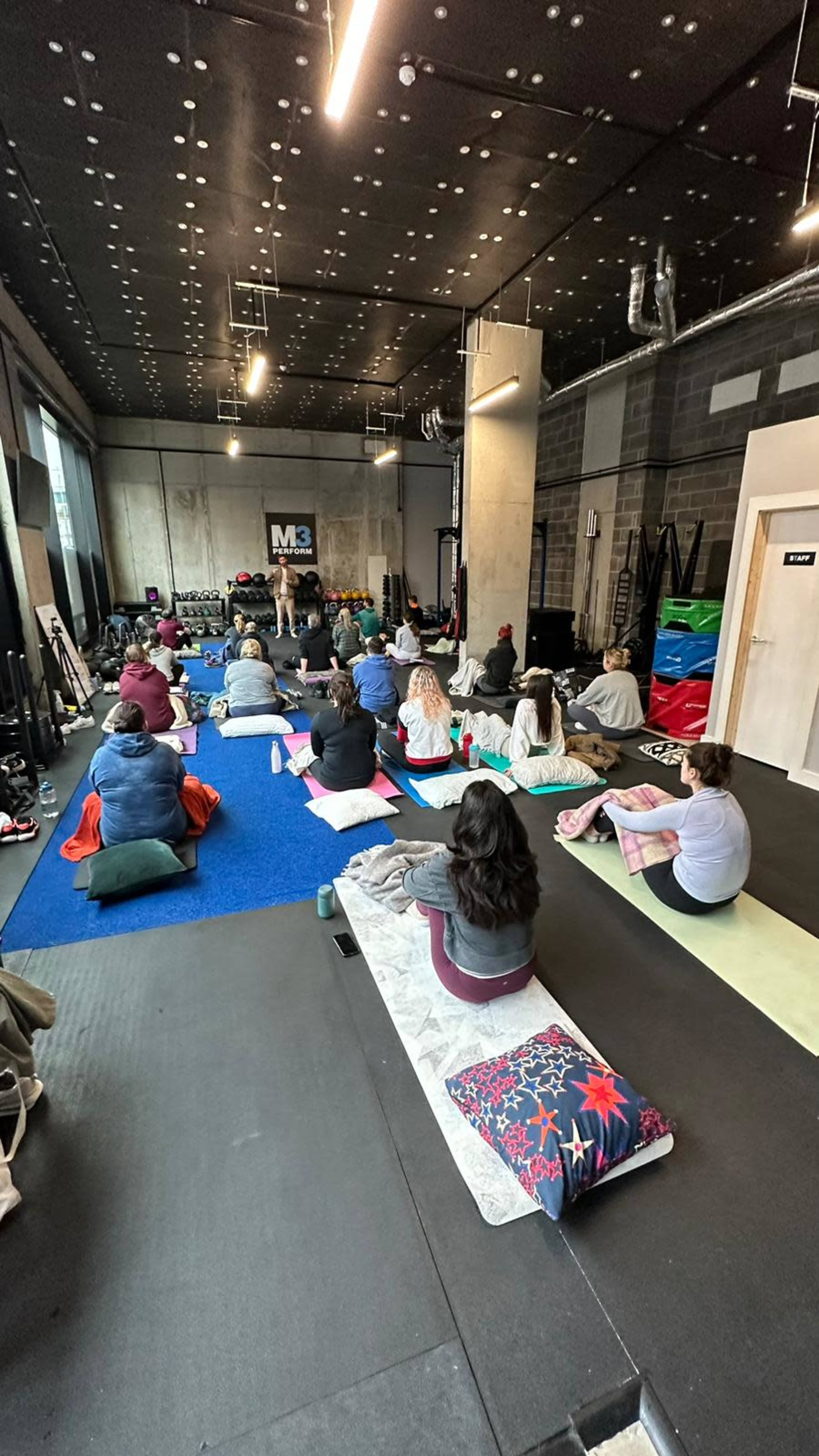 A group of people is seated on mats in a fitness studio, facing an instructor who is demonstrating a position.