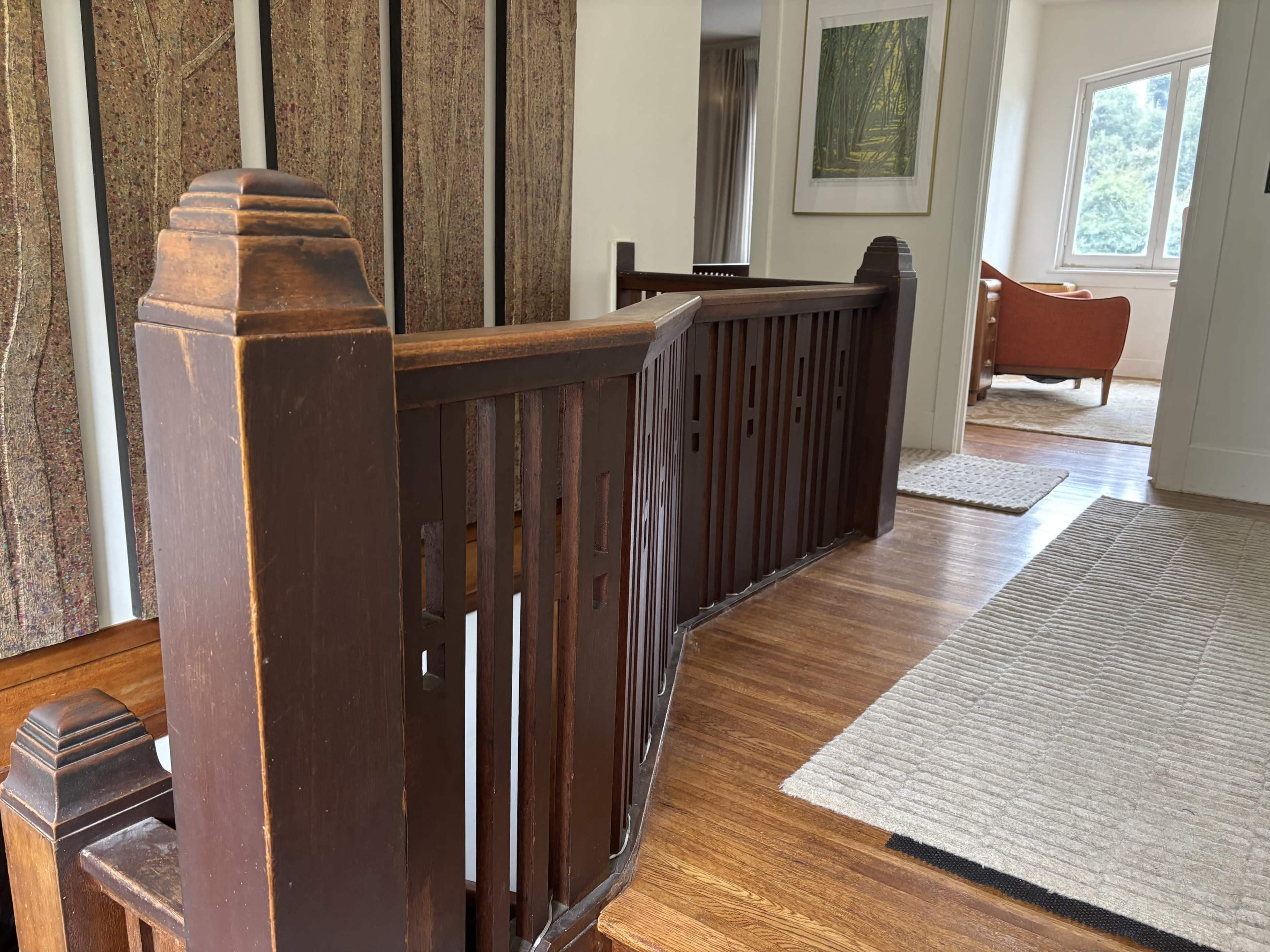 The image shows a wooden railing and partition in a hallway, leading toward a light-filled room with a chair and rug.