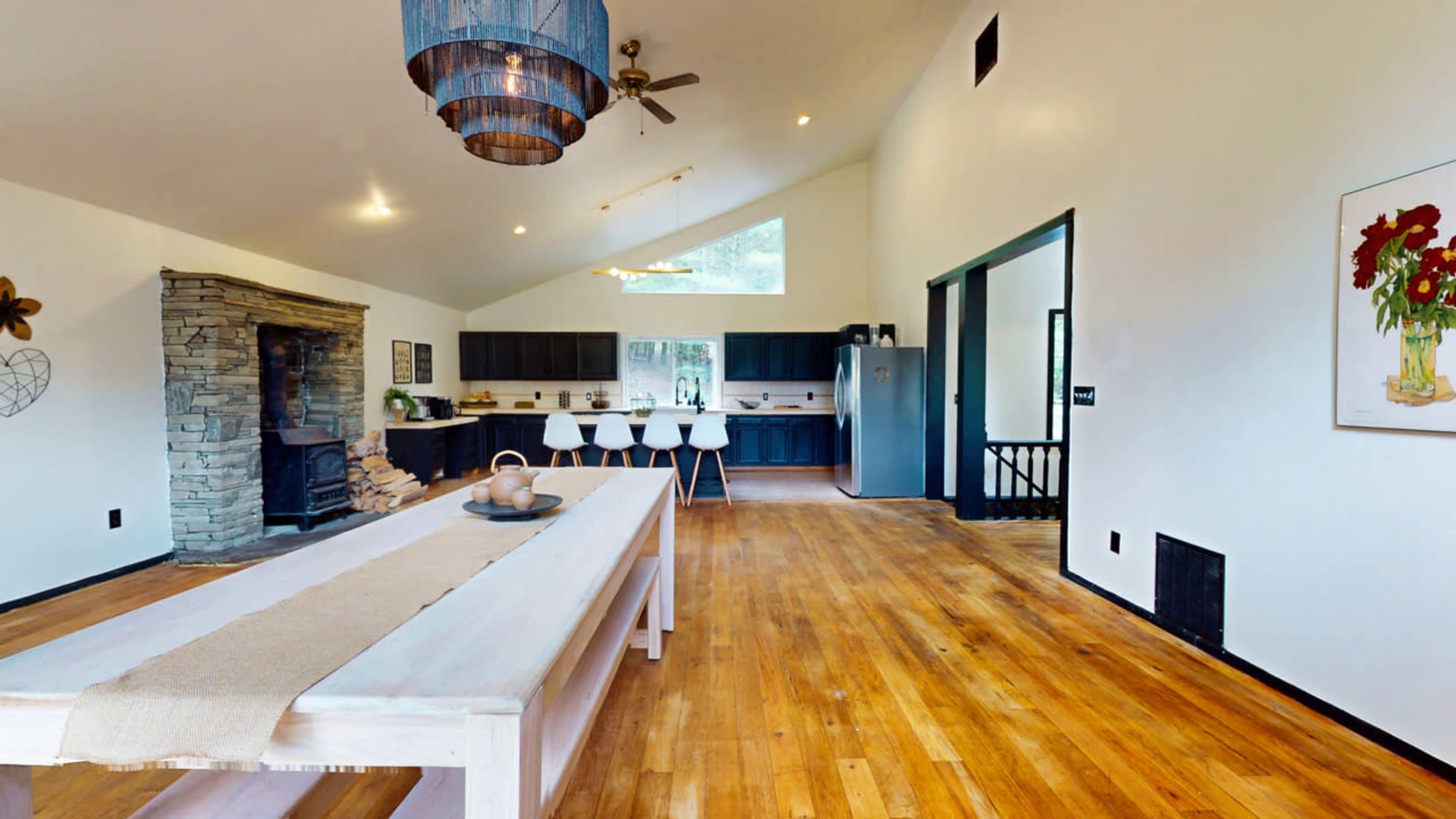 A modern kitchen with a wooden dining table, a stone fireplace, and dark cabinetry complemented by white walls and natural wood flooring.