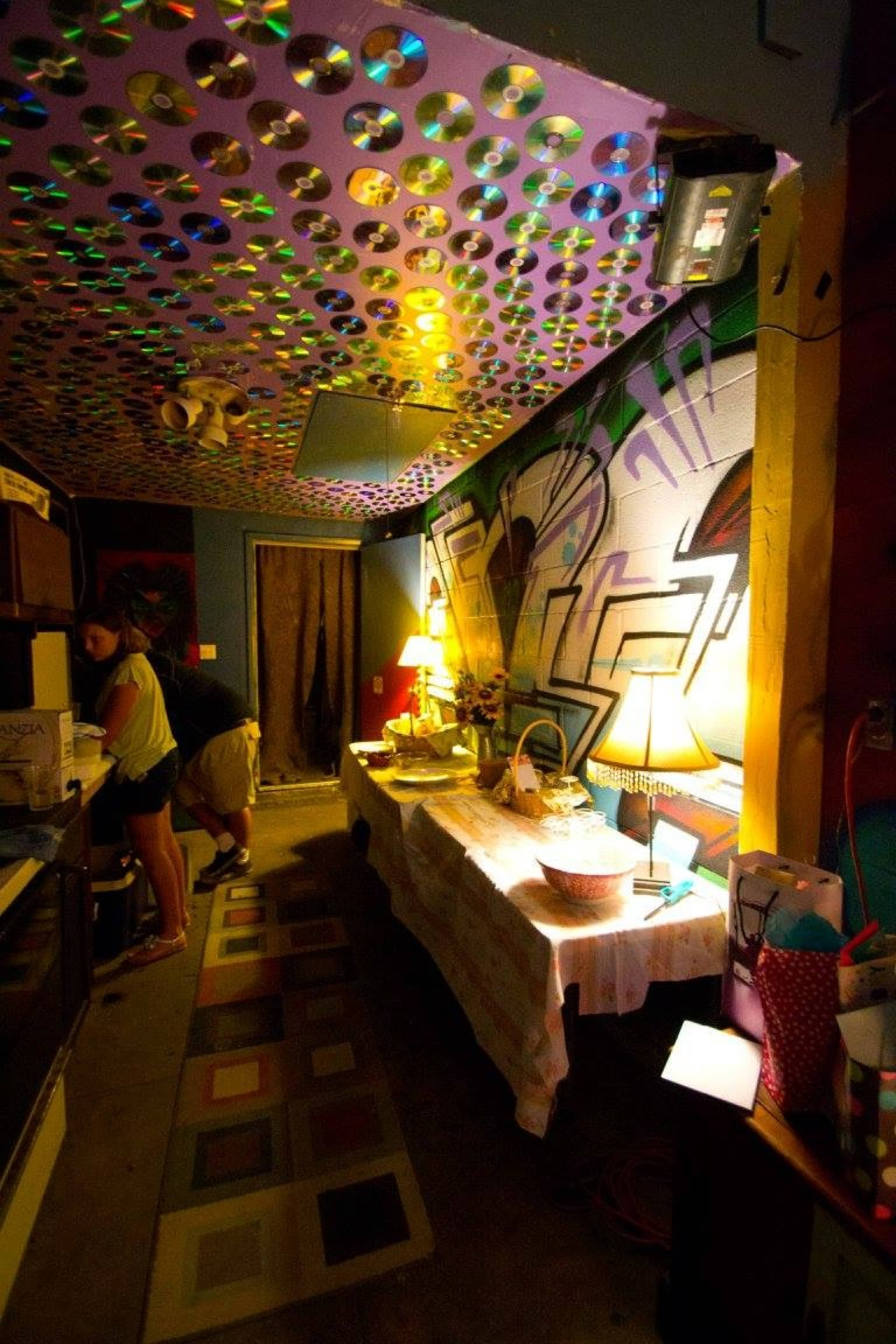 A small kitchen area features a ceiling adorned with colorful CDs, a table set with various items, and a person preparing food at the counter.