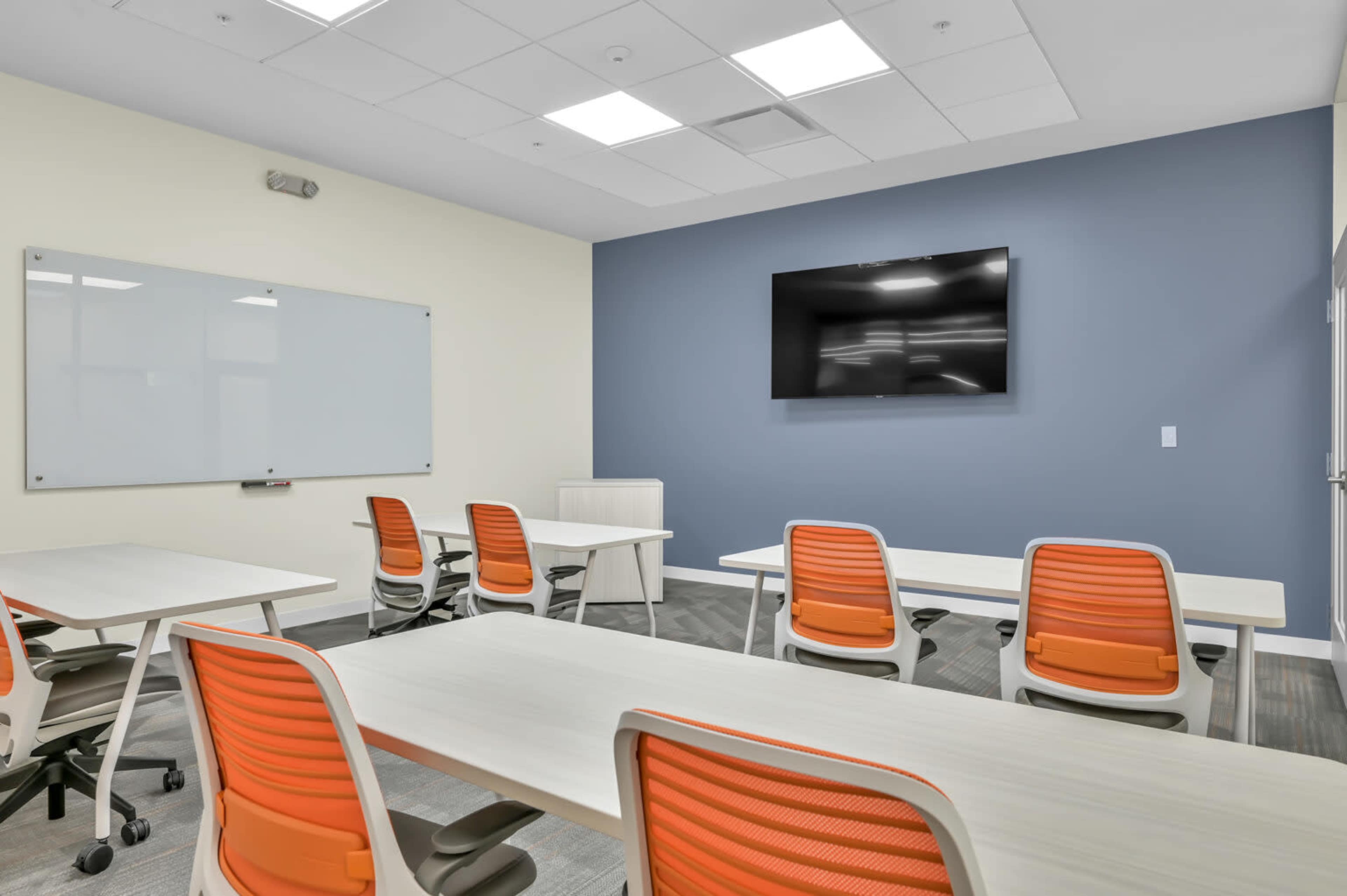A modern conference room features several tables and chairs with orange accents, a whiteboard, and a wall-mounted television.